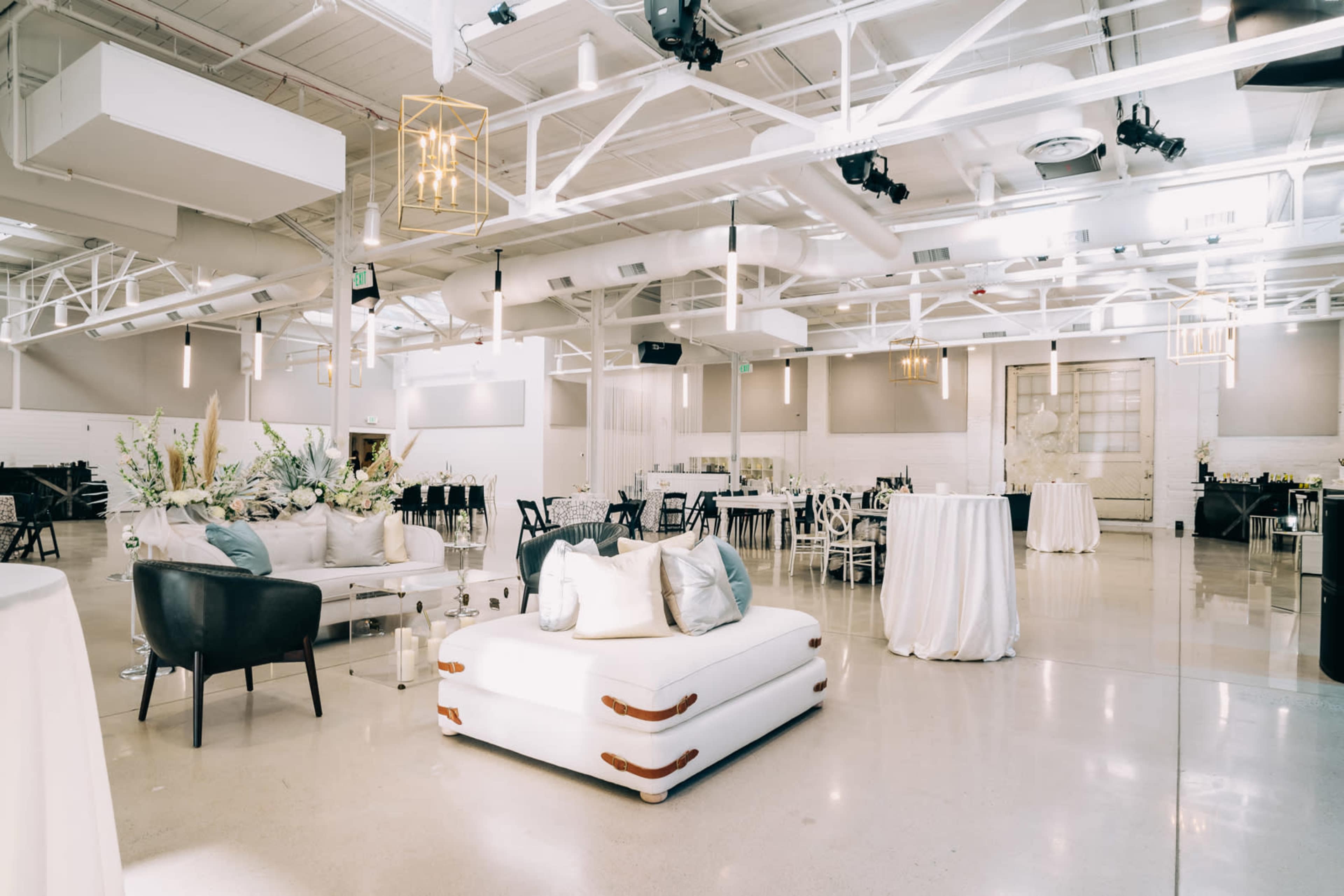 A spacious event venue featuring modern decor, white seating areas, and multiple tables set up for an event.