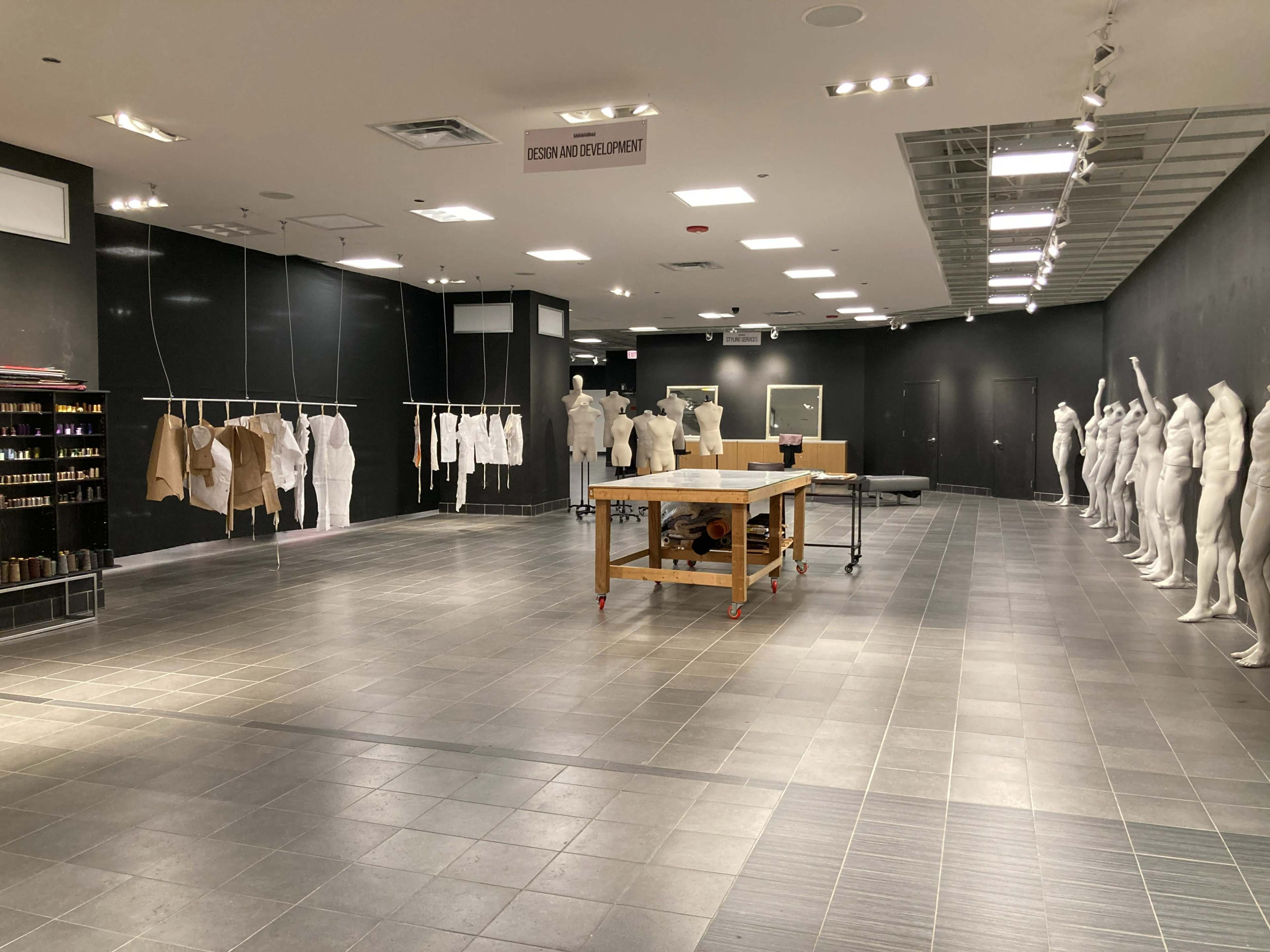 The image shows a spacious, empty room with black walls, featuring several mannequins dressed in neutral clothing, hanging garments, and a central worktable.