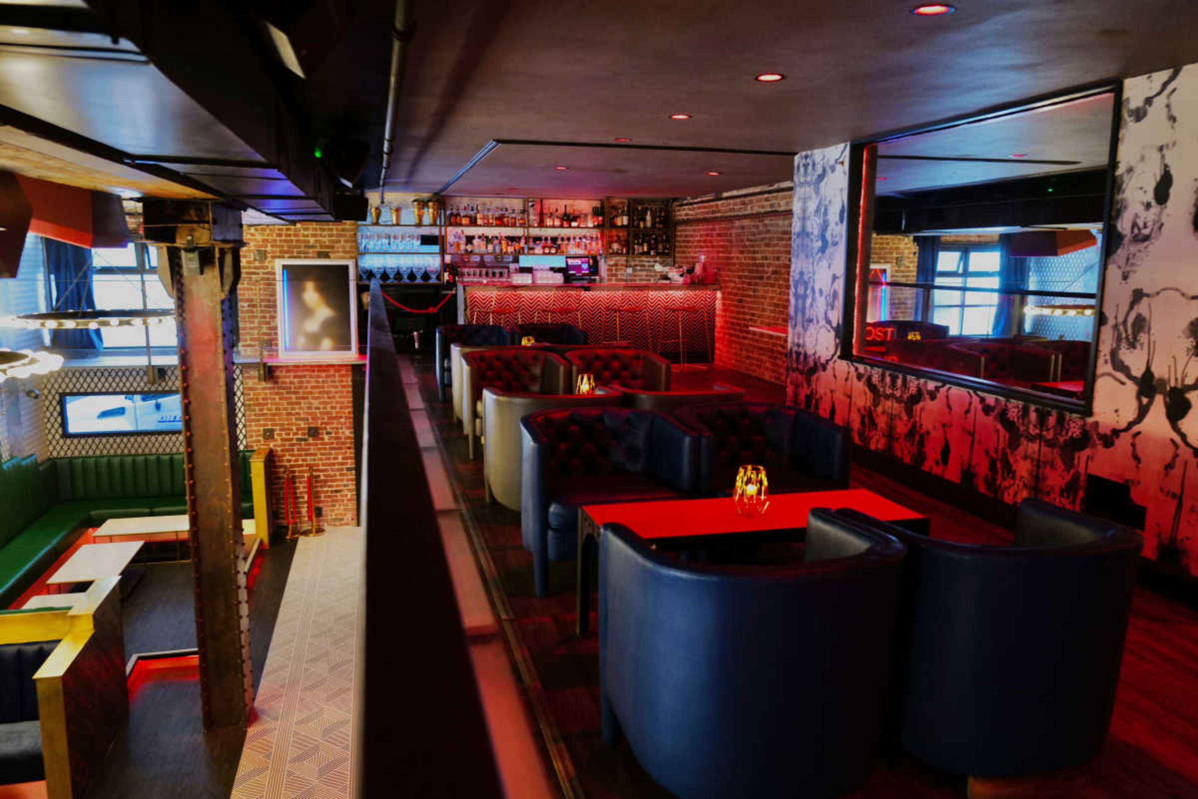 The image shows a dimly lit bar with a modern interior featuring plush seating areas and a long bar counter illuminated by ambient lighting.