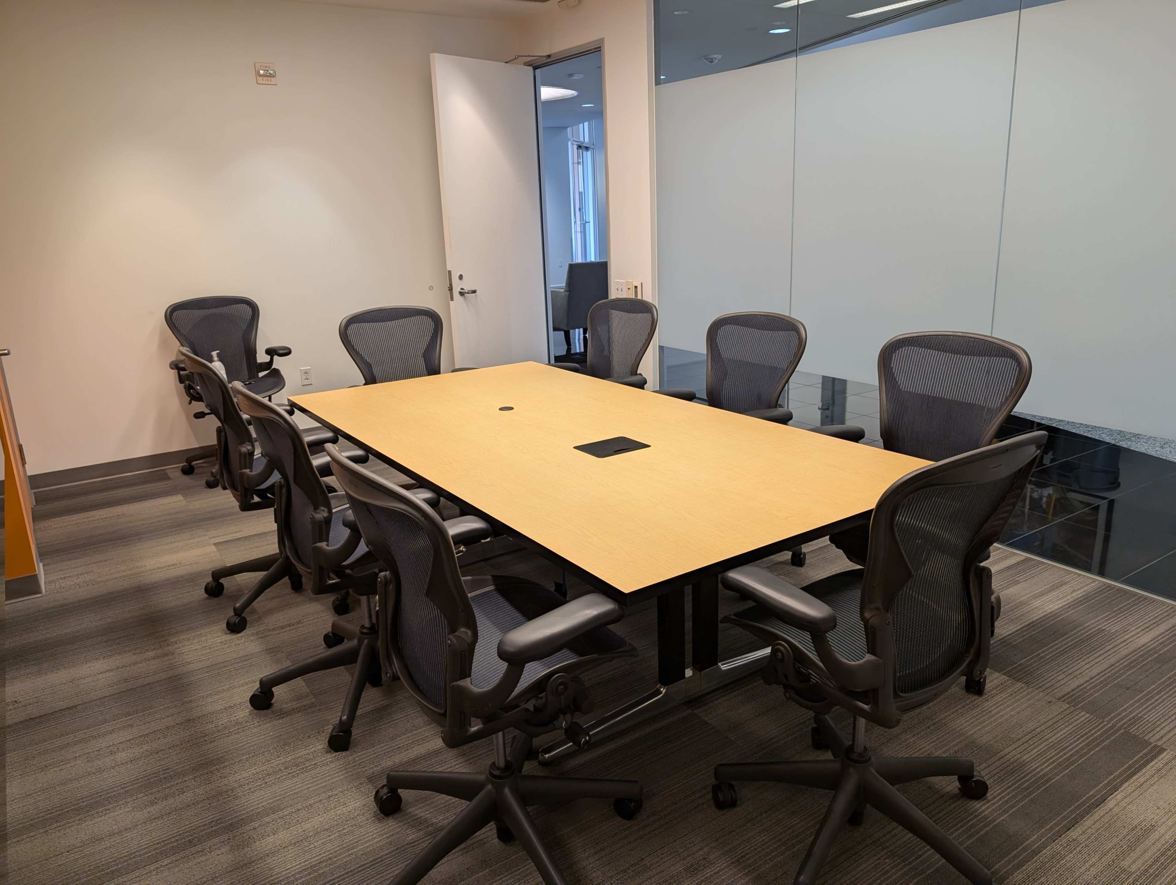 A modern conference room features a wooden table surrounded by ergonomic chairs, with glass walls and ample natural light.