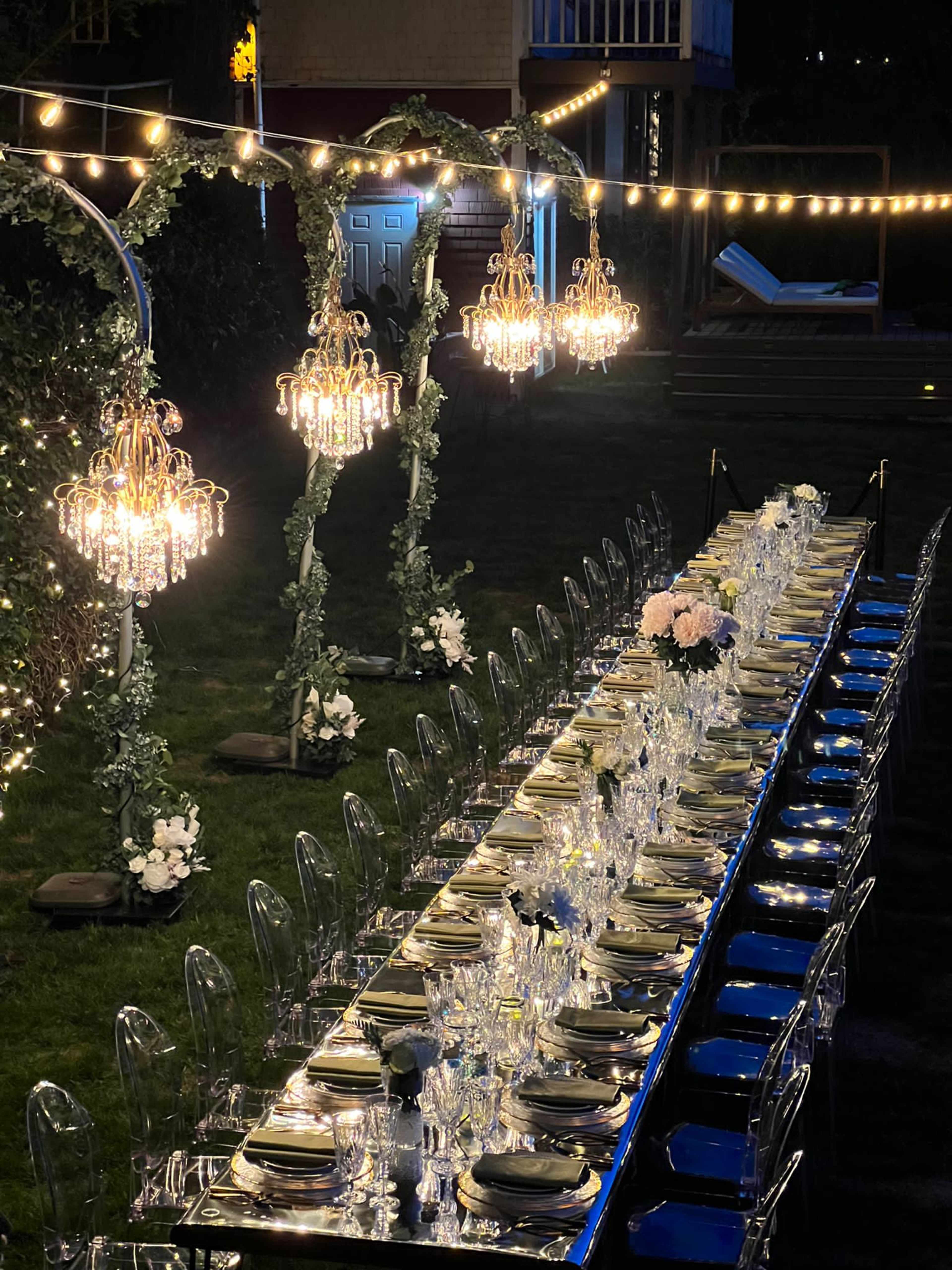 A long, elegantly set dining table with crystal glassware and floral centerpieces is illuminated by hanging chandeliers and string lights in a garden setting.