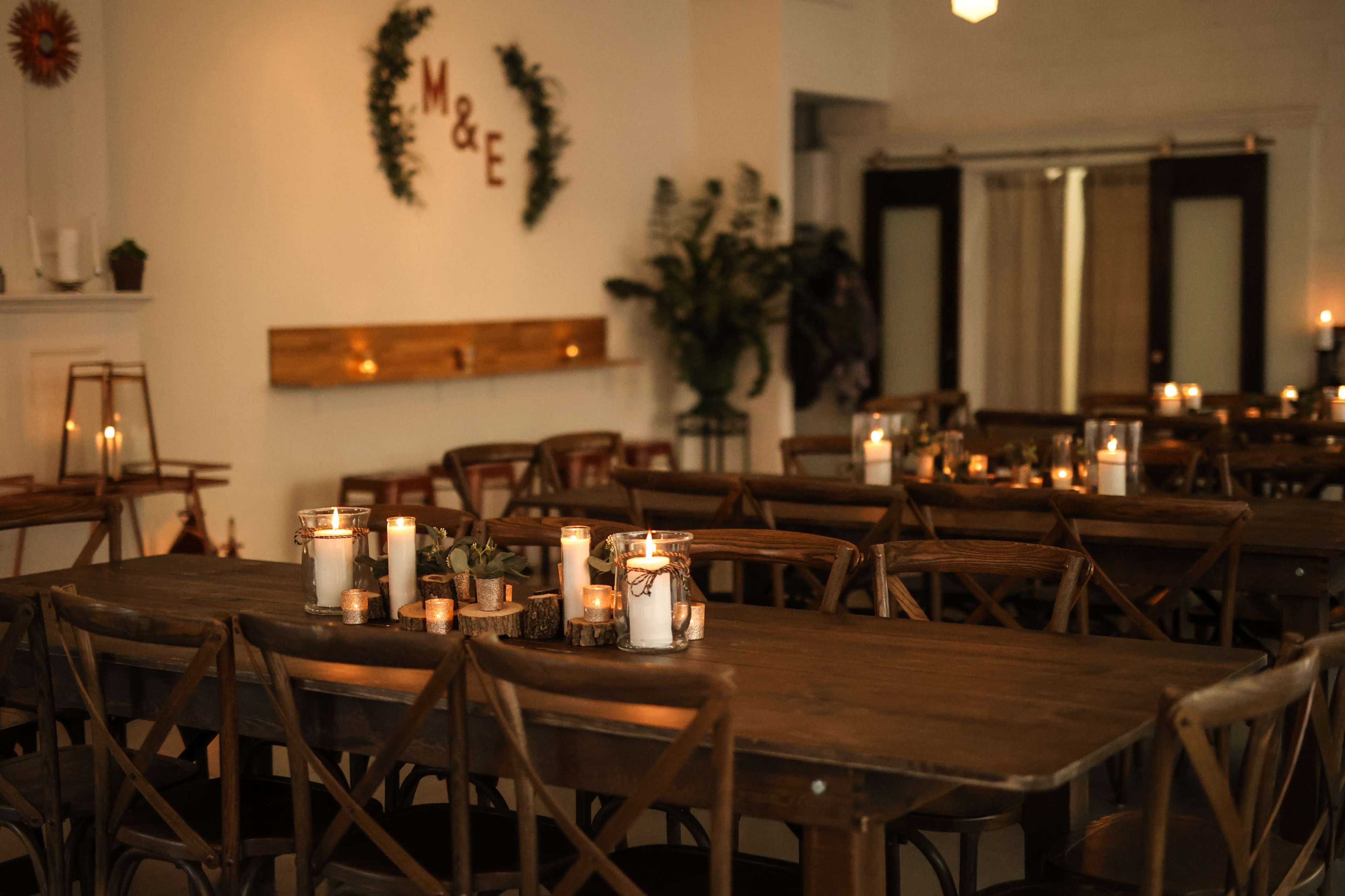 The image shows a warmly lit event space with wooden tables arranged for seating, adorned with candles and simple decorations.