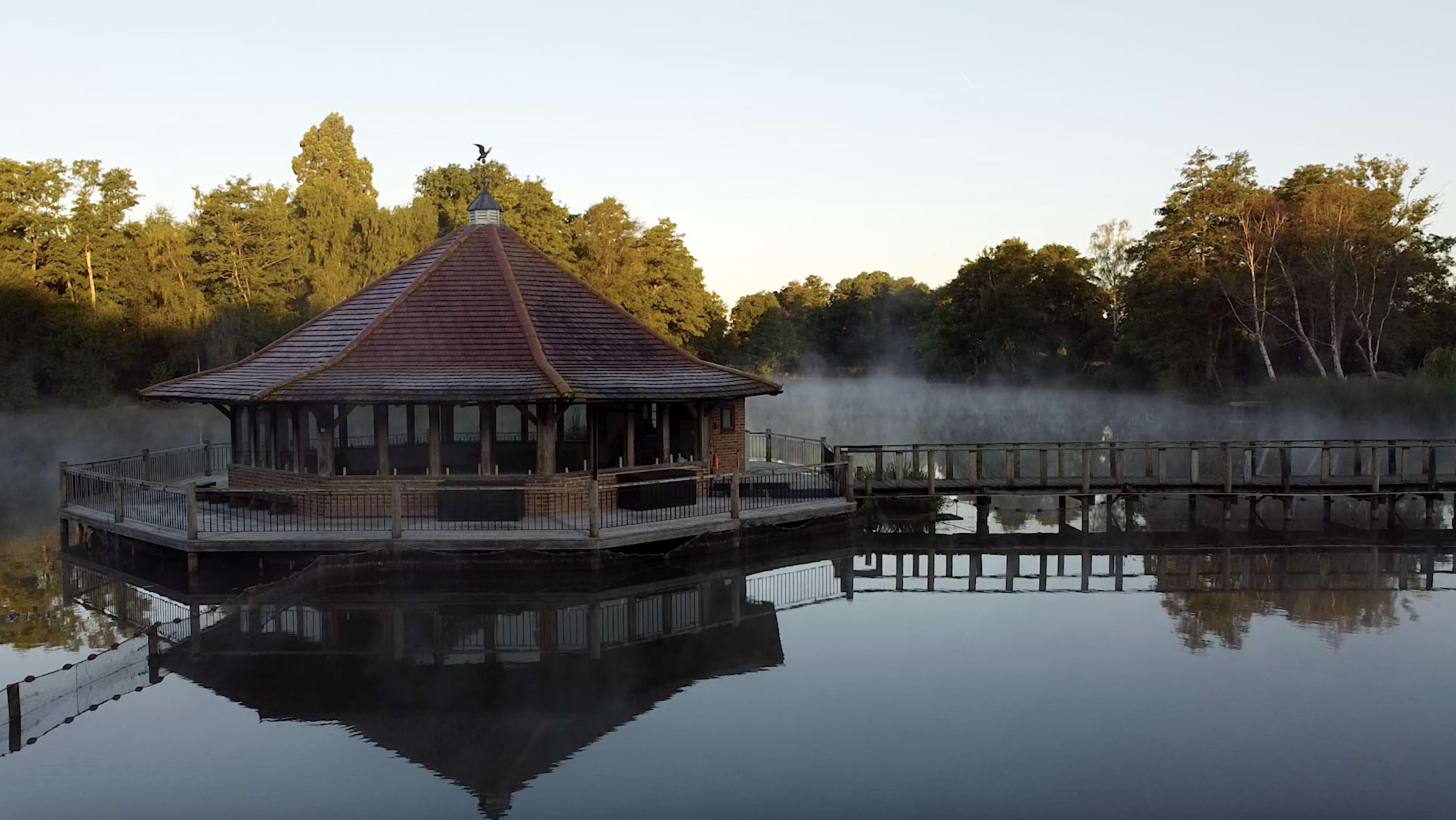 A wooden pavilion with a conical roof is situated on a misty lake, reflecting in the calm water.