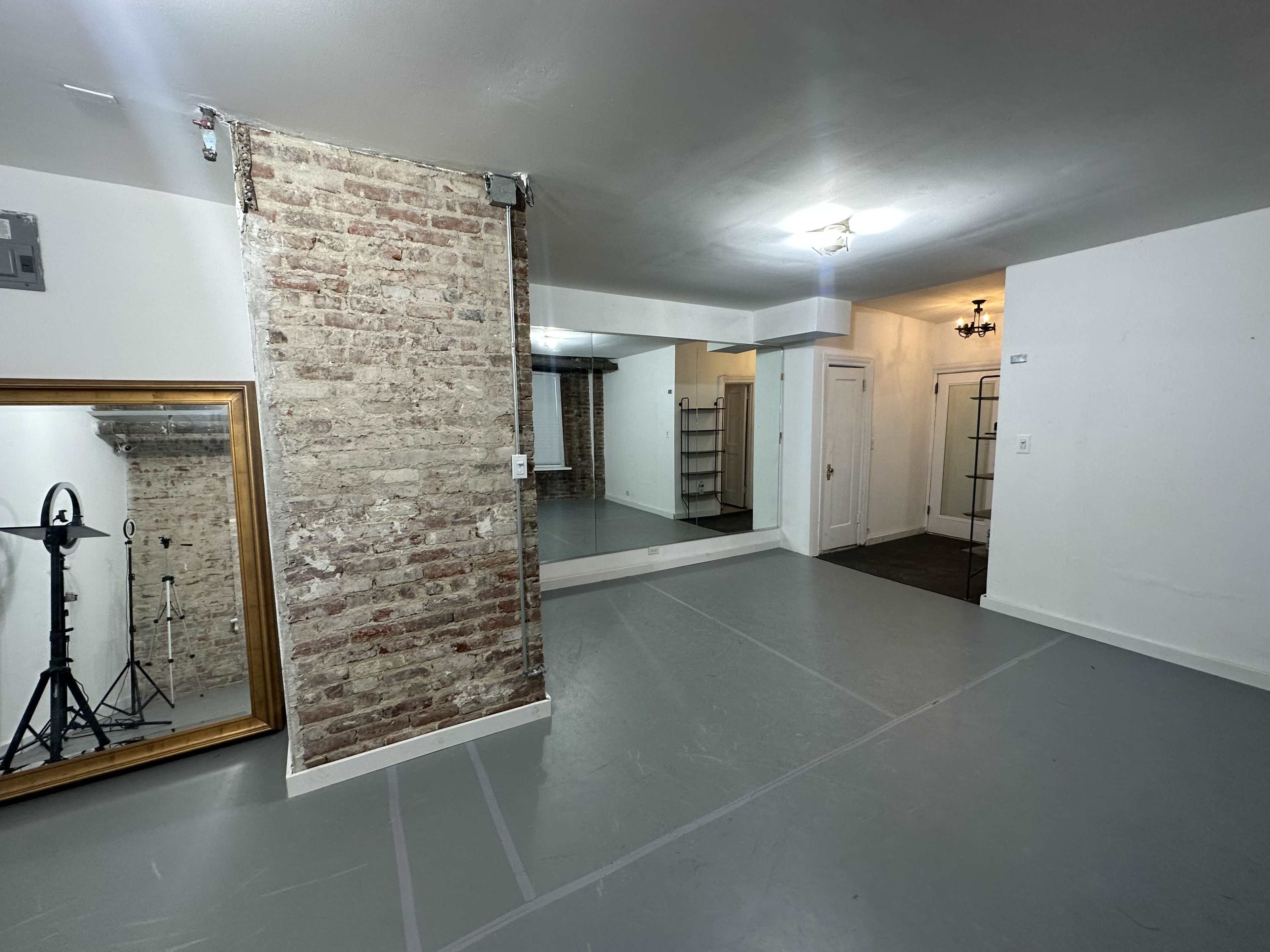 A spacious room with exposed brick walls, a large mirror, and a sectioned area featuring a standing light and shelves.
