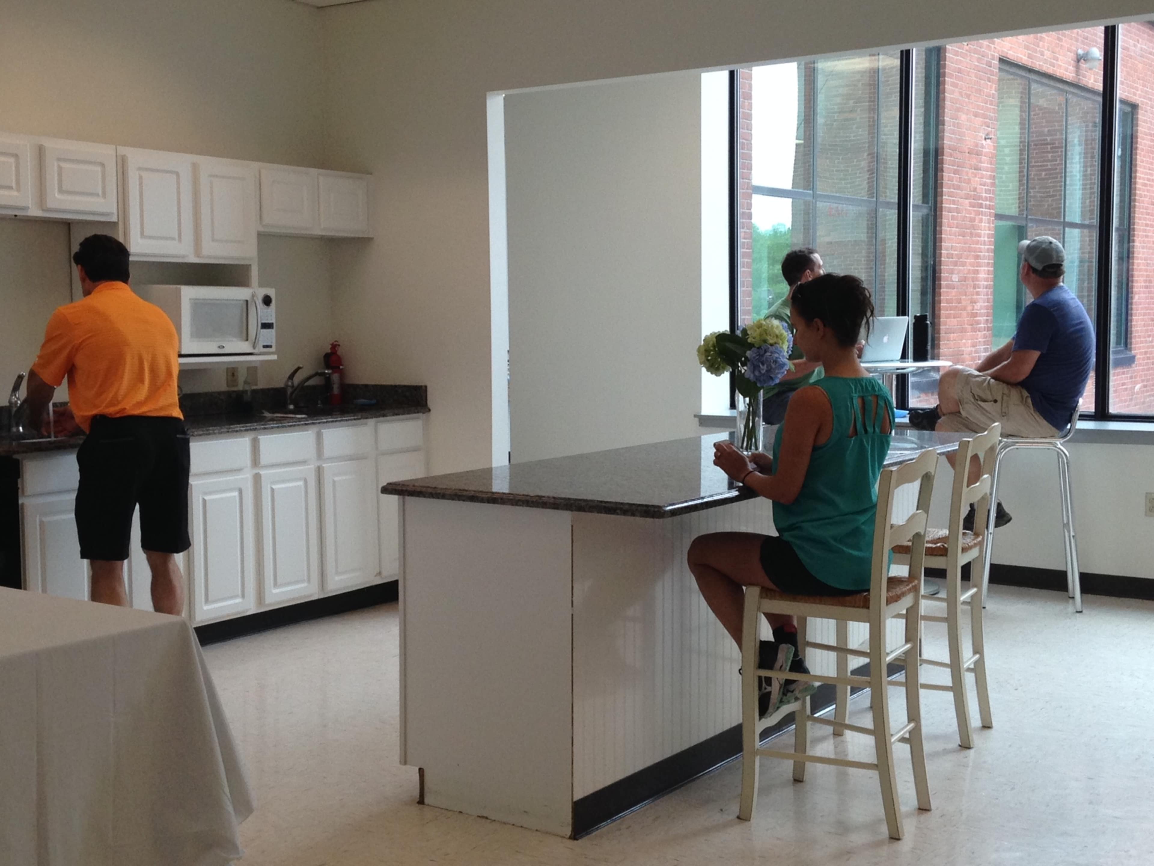 A kitchen area features a person washing dishes, while another sits at a counter and two people work on laptops at a table.