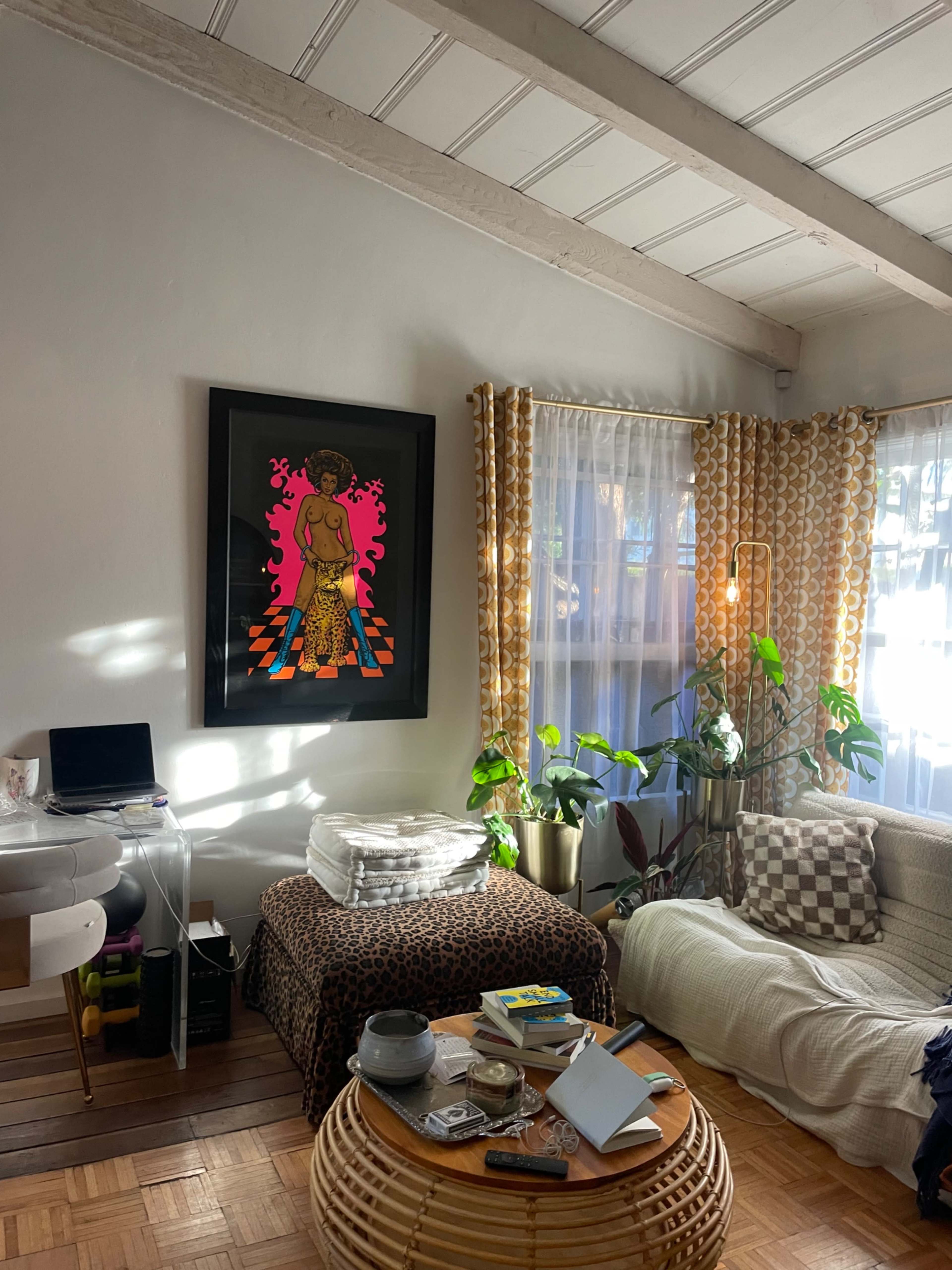 The living room features a light-colored sofa, a patterned table, a large framed artwork on the wall, and various houseplants near a window with sheer curtains.