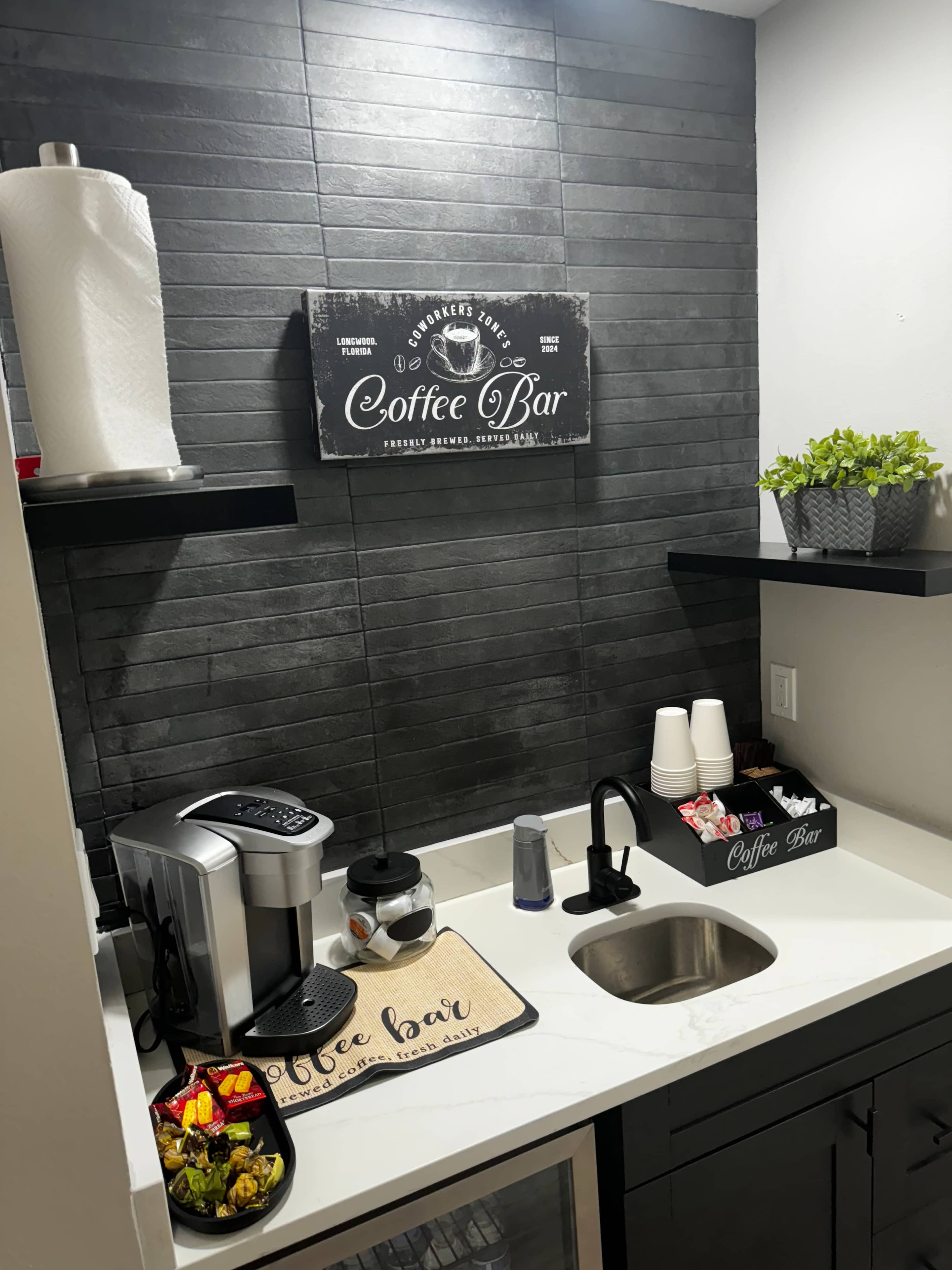 The image shows a small coffee station with a coffee machine, sink, and decorative items on shelves in a dark-themed space.