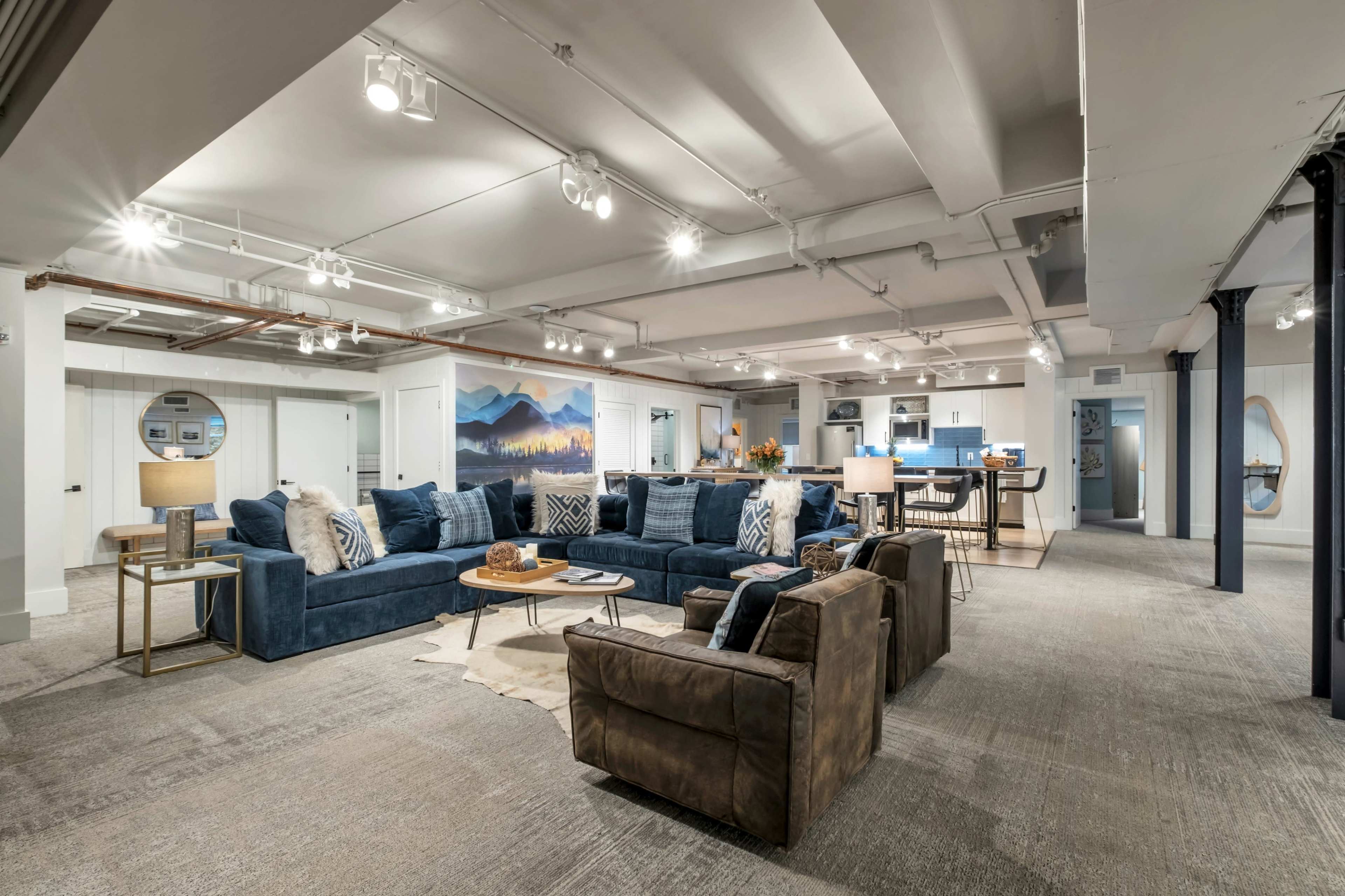 The image shows a spacious, modern basement living area with a seating arrangement featuring blue couches and a brown armchair, accented by a large mural of mountains and a well-lit open kitchen in the background.