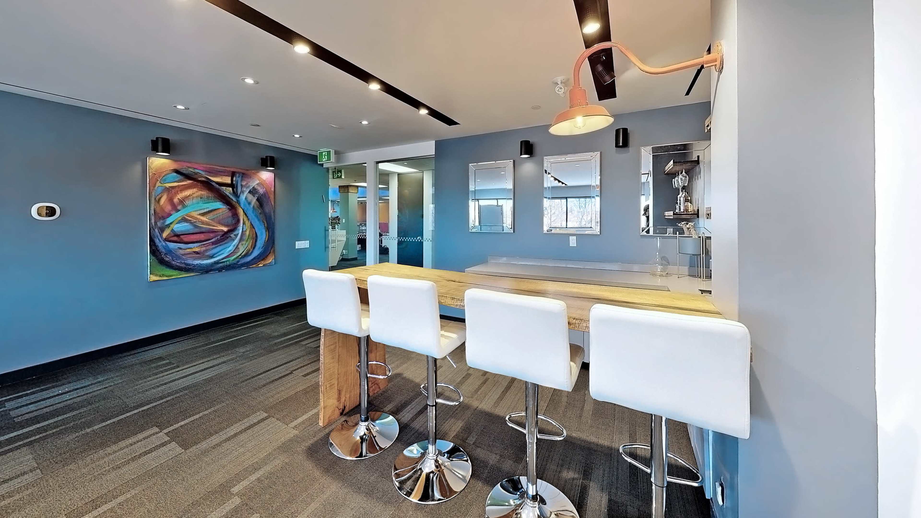 The image shows a modern, minimalist bar area with four white stools surrounding a wooden counter, set against a blue wall featuring colorful artwork and several mirrors.