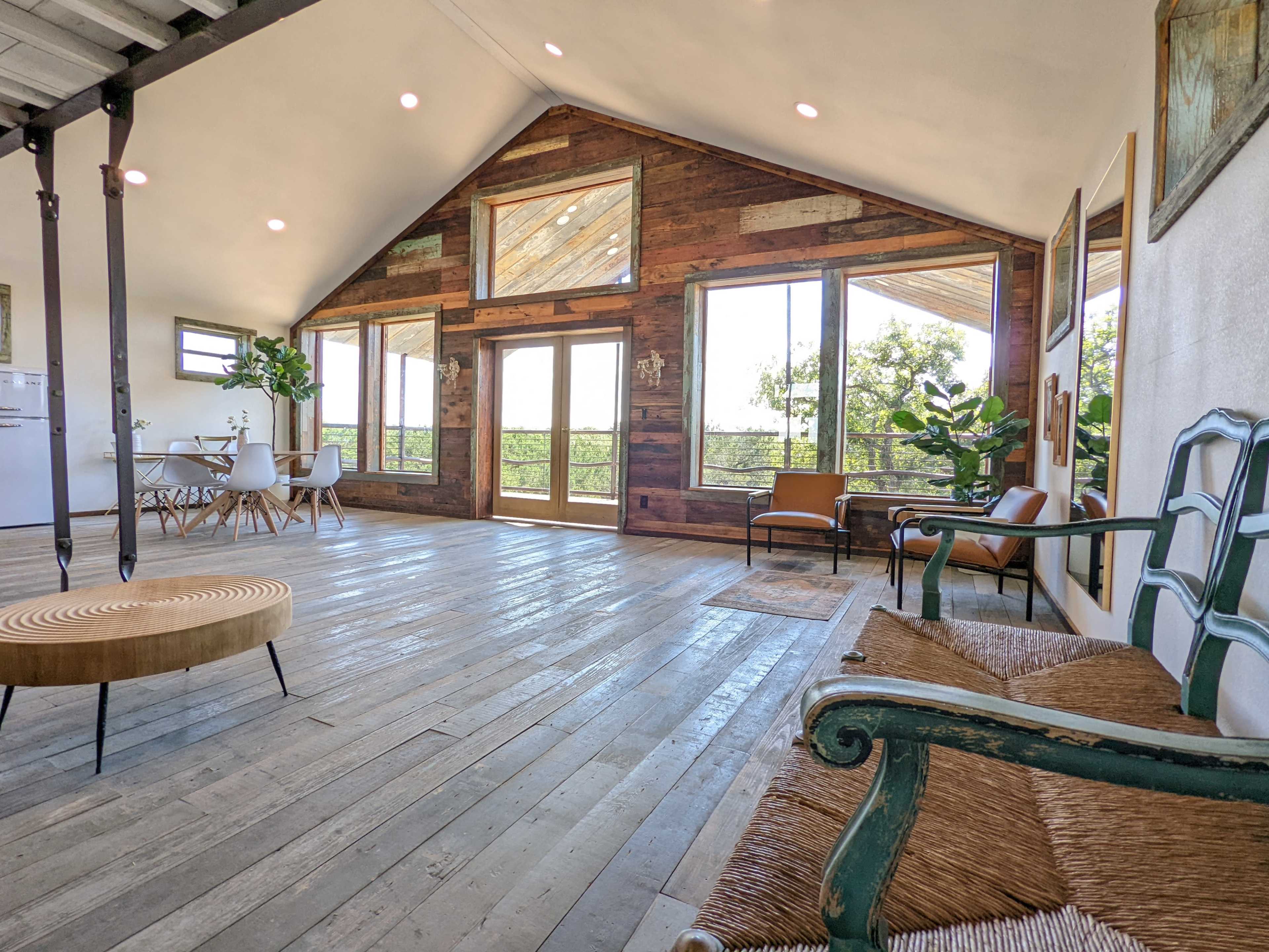 A spacious, modern interior with wooden floors, large windows, and a mix of seating options including chairs and a round ottoman.