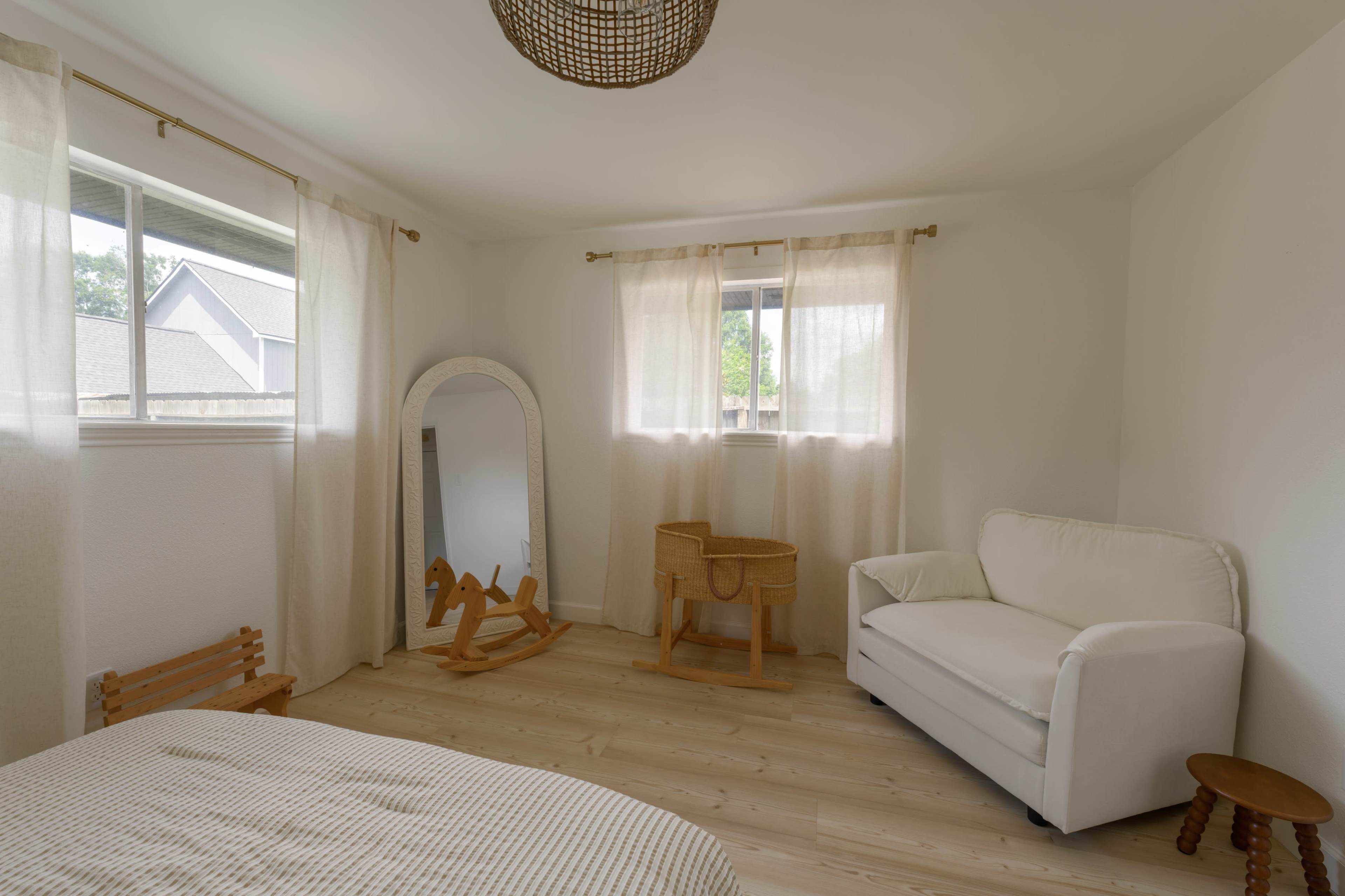 The room features a white couch, a large mirror, a wooden rocking horse, and a set of wicker baskets, all set against light-colored walls and wooden flooring.