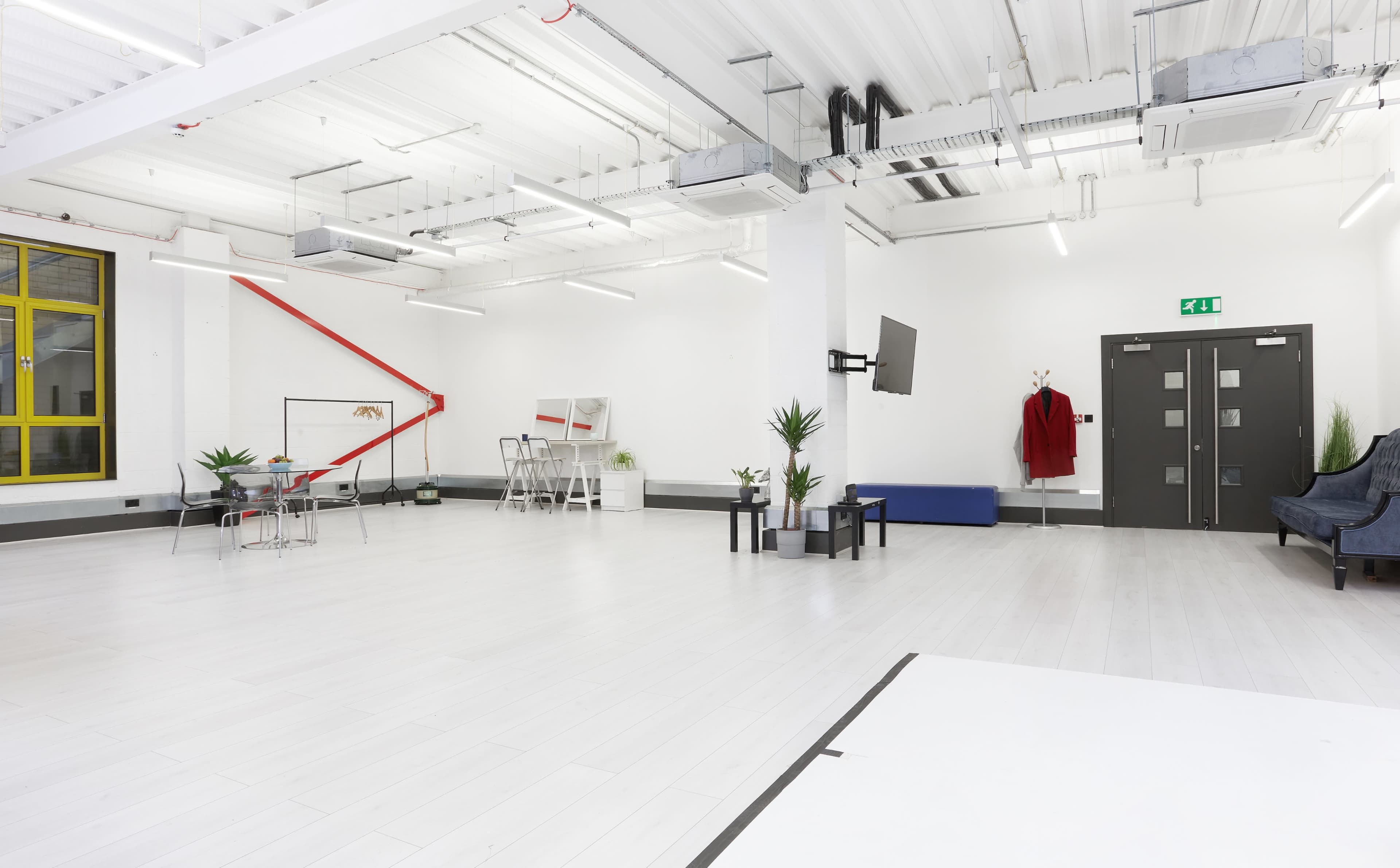 A spacious, well-lit studio featuring white walls, a seating area with a blue chair, a table set with chairs, and a red coat hanging on a wall.