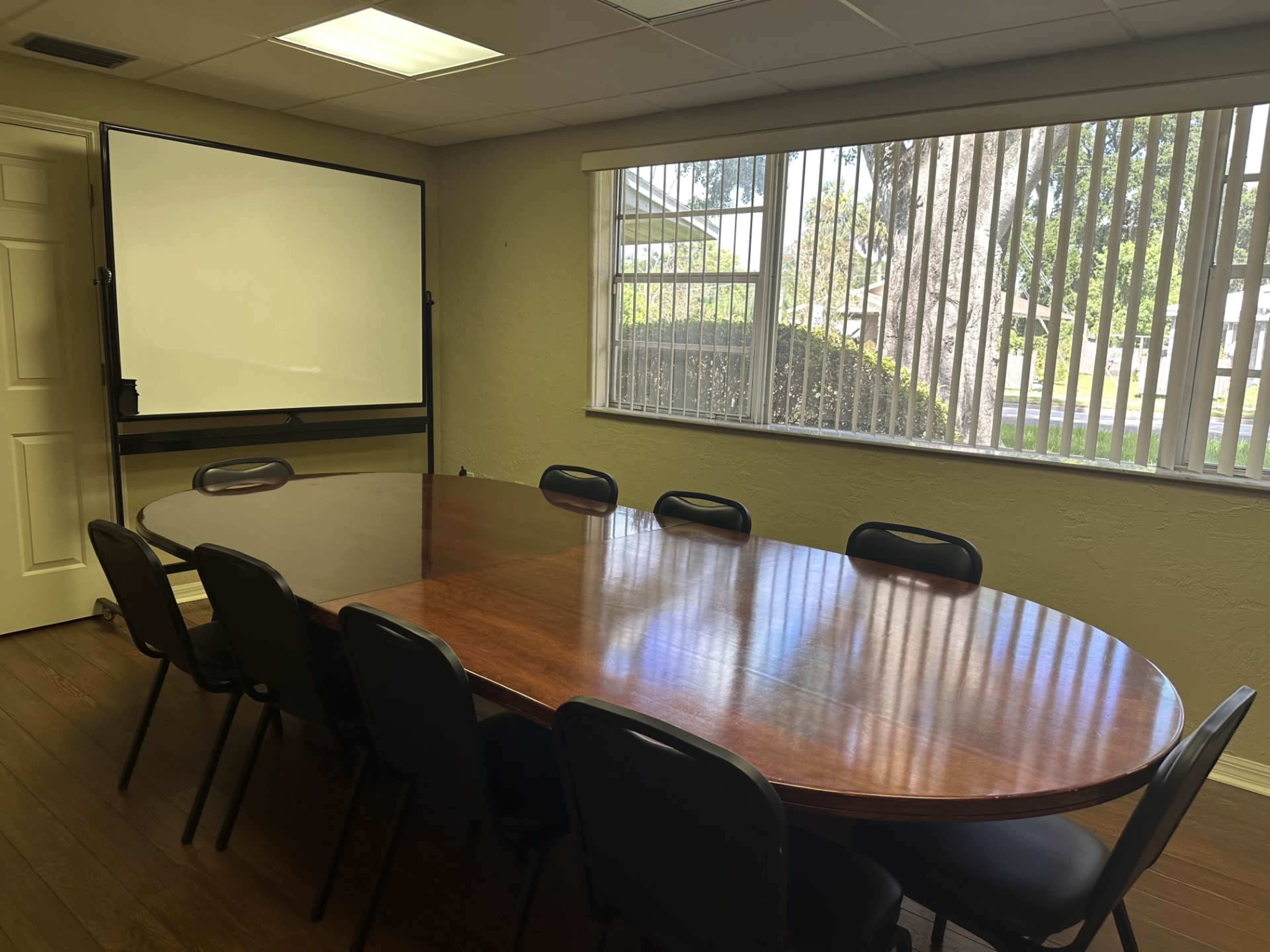 A large, oval wooden conference table is surrounded by black chairs in a well-lit meeting room with a whiteboard and windows displaying greenery outside.
