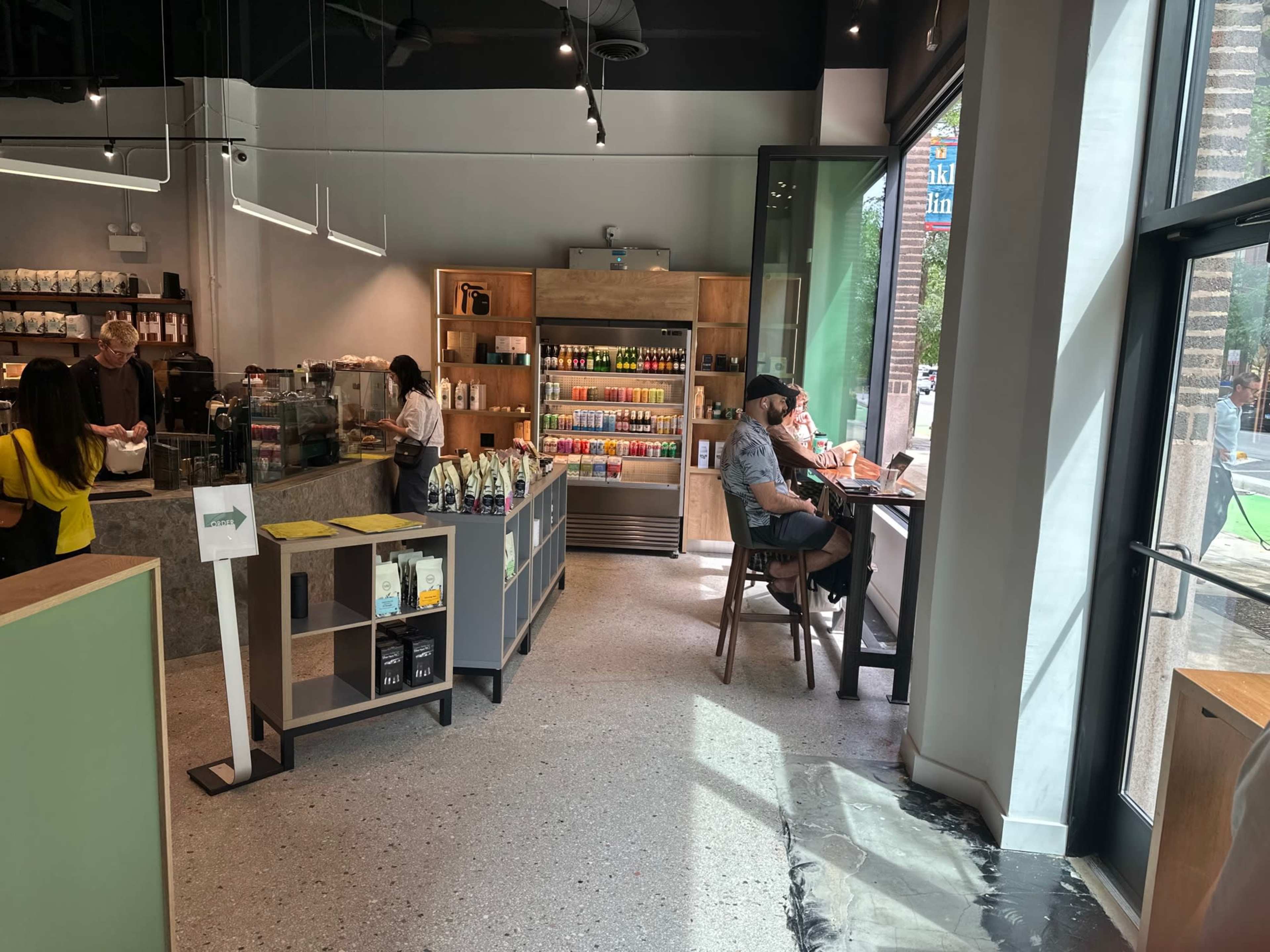 The interior of a café with a coffee counter, shelves lined with beverages, and customers seated at tables near large windows.