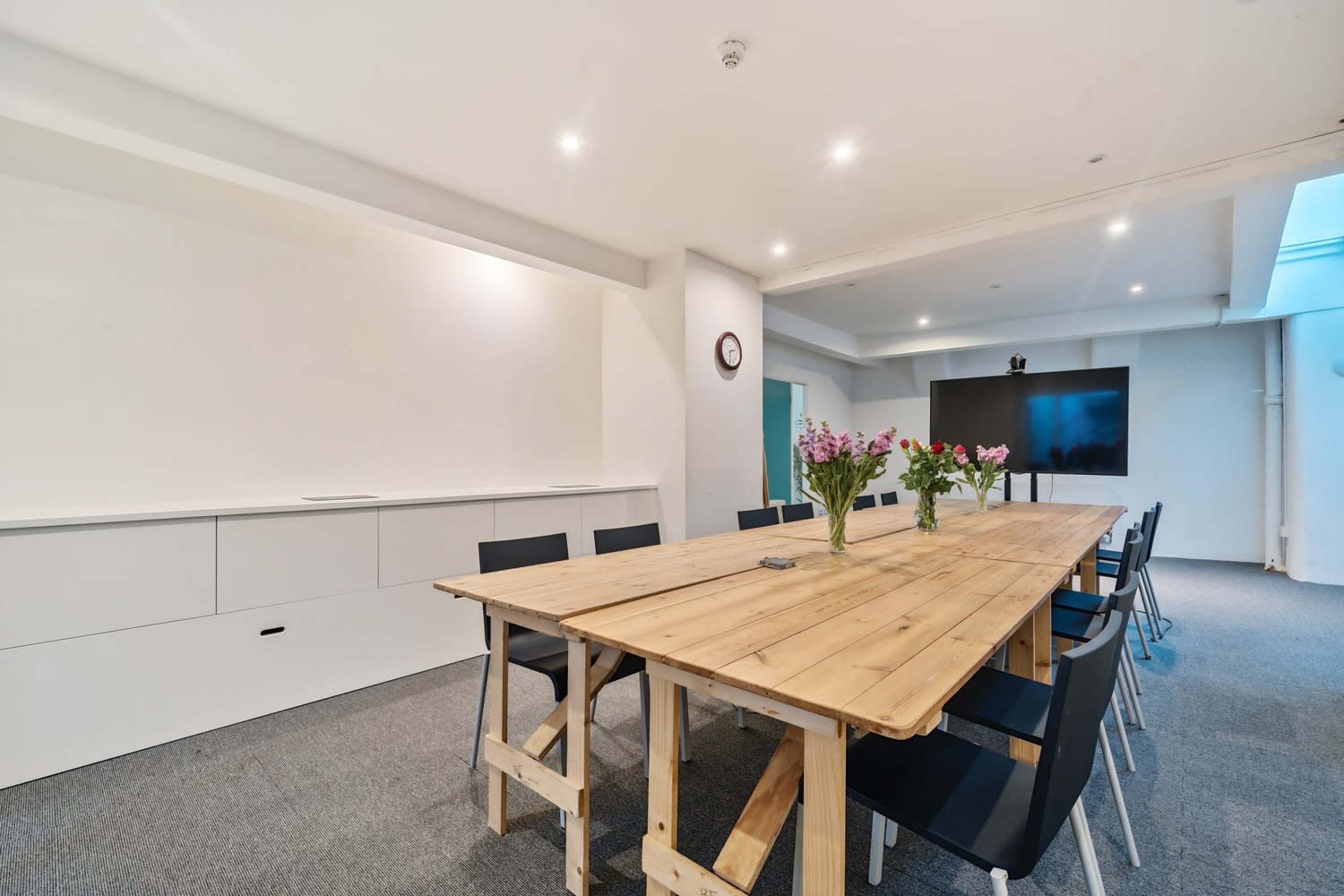 A large wooden table surrounded by black chairs is set up in a modern conference room with a television and wall-mounted clock.