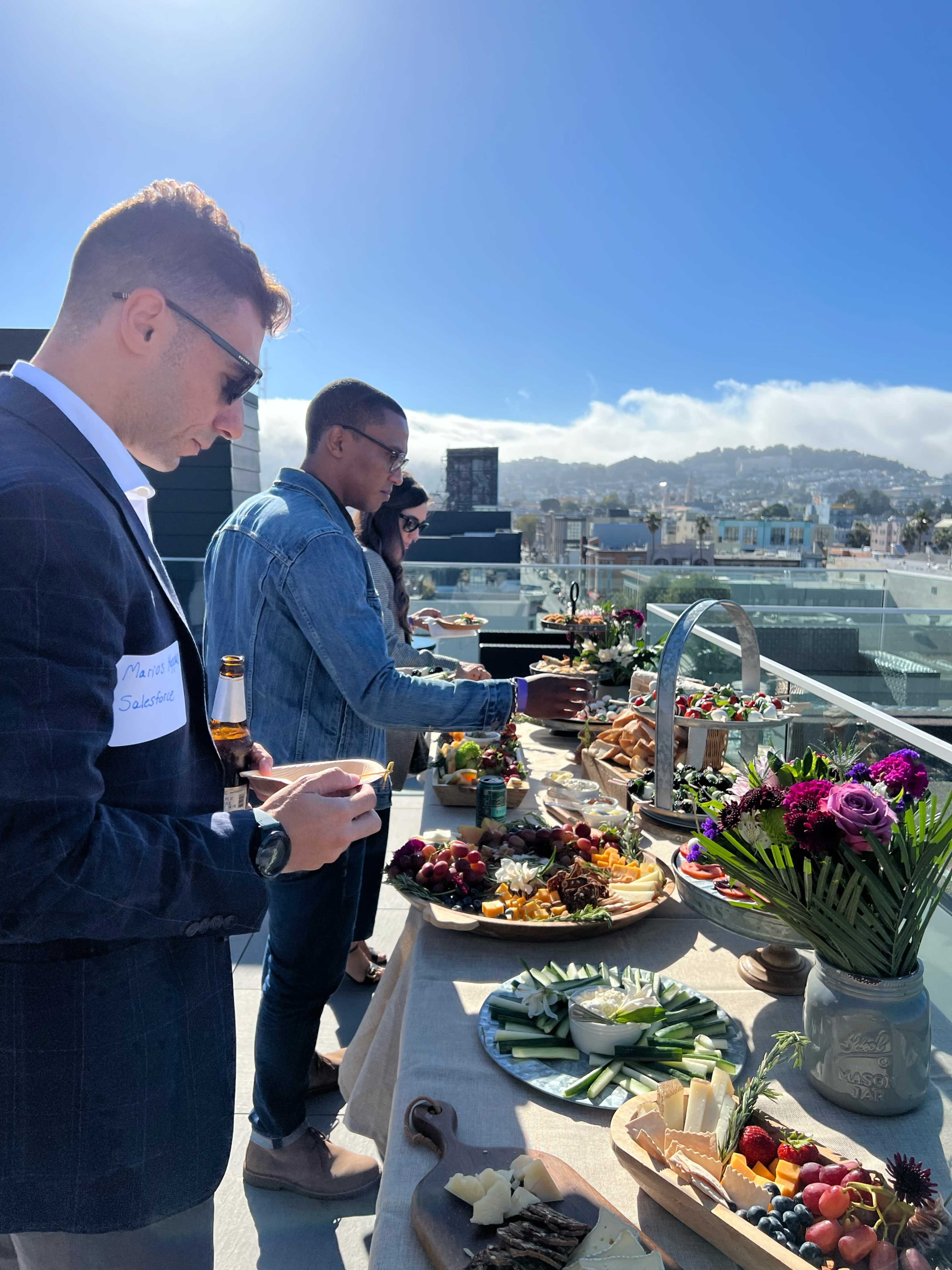 A group of people is enjoying a buffet spread of food and drinks on a rooftop with a city view.