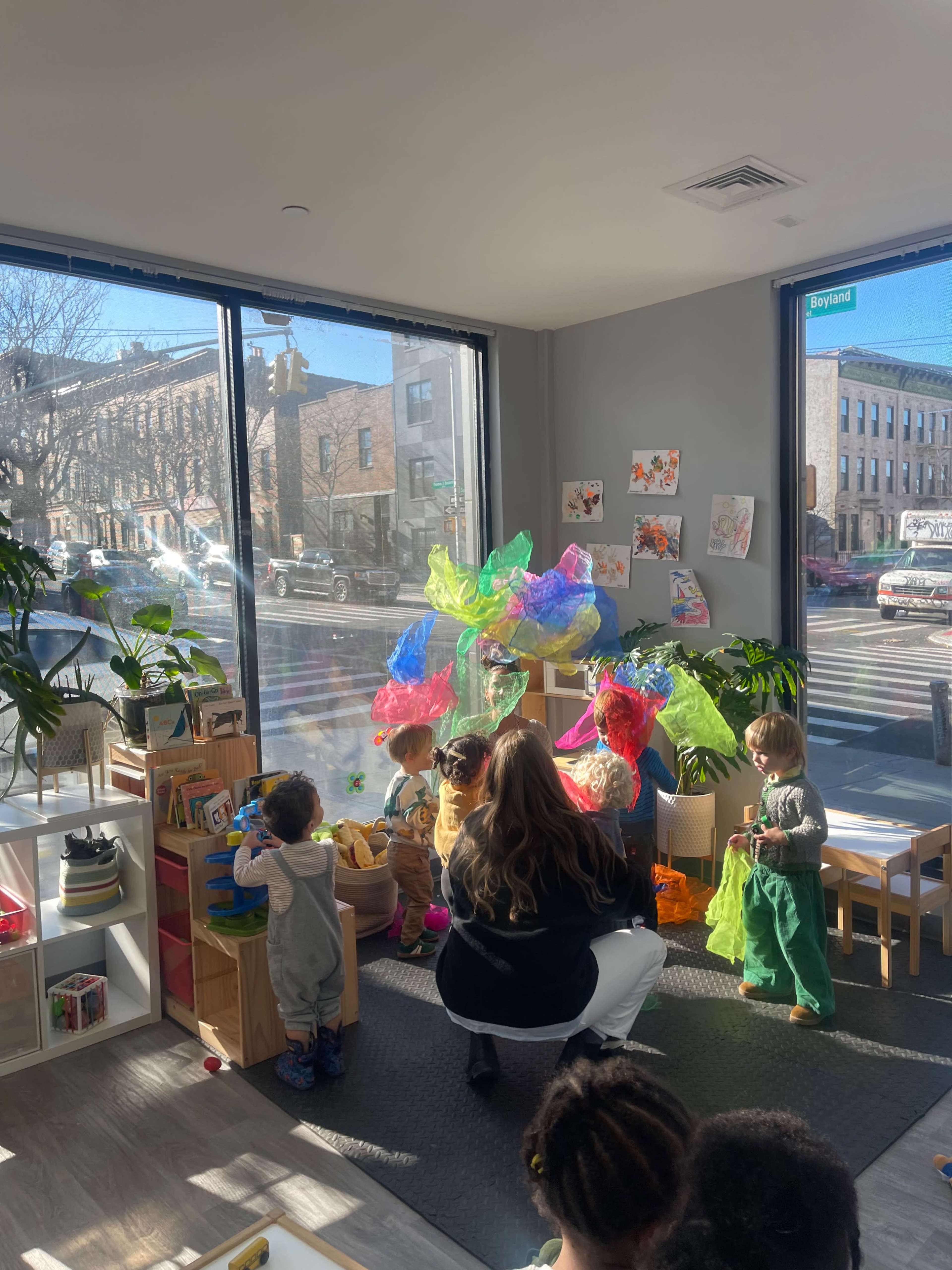 A group of children play with colorful materials in a bright room filled with plants and large windows overlooking a street.