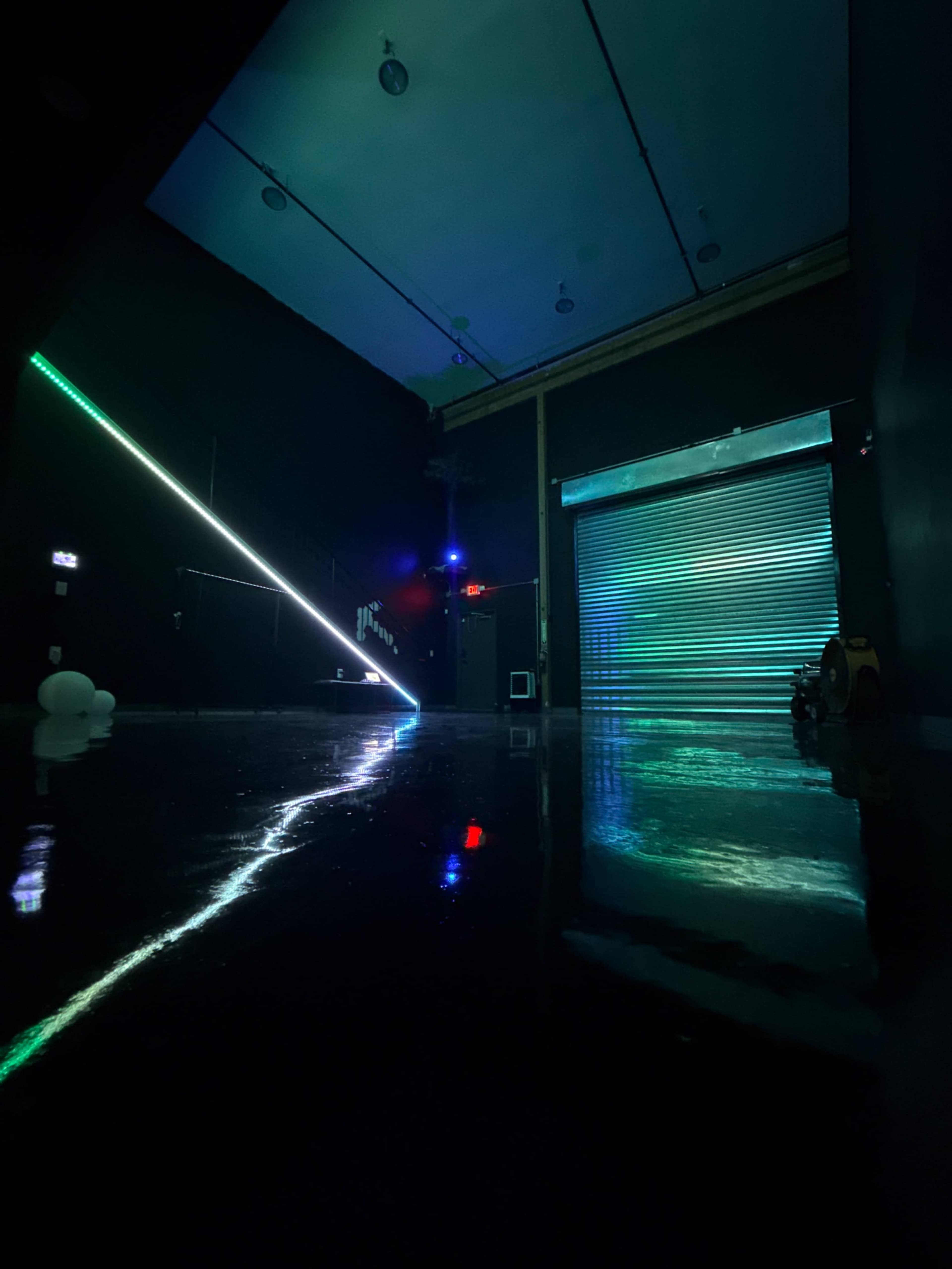 The image shows a dimly lit room with reflective black floors, featuring neon lights creating geometric patterns and a closed metallic garage door.