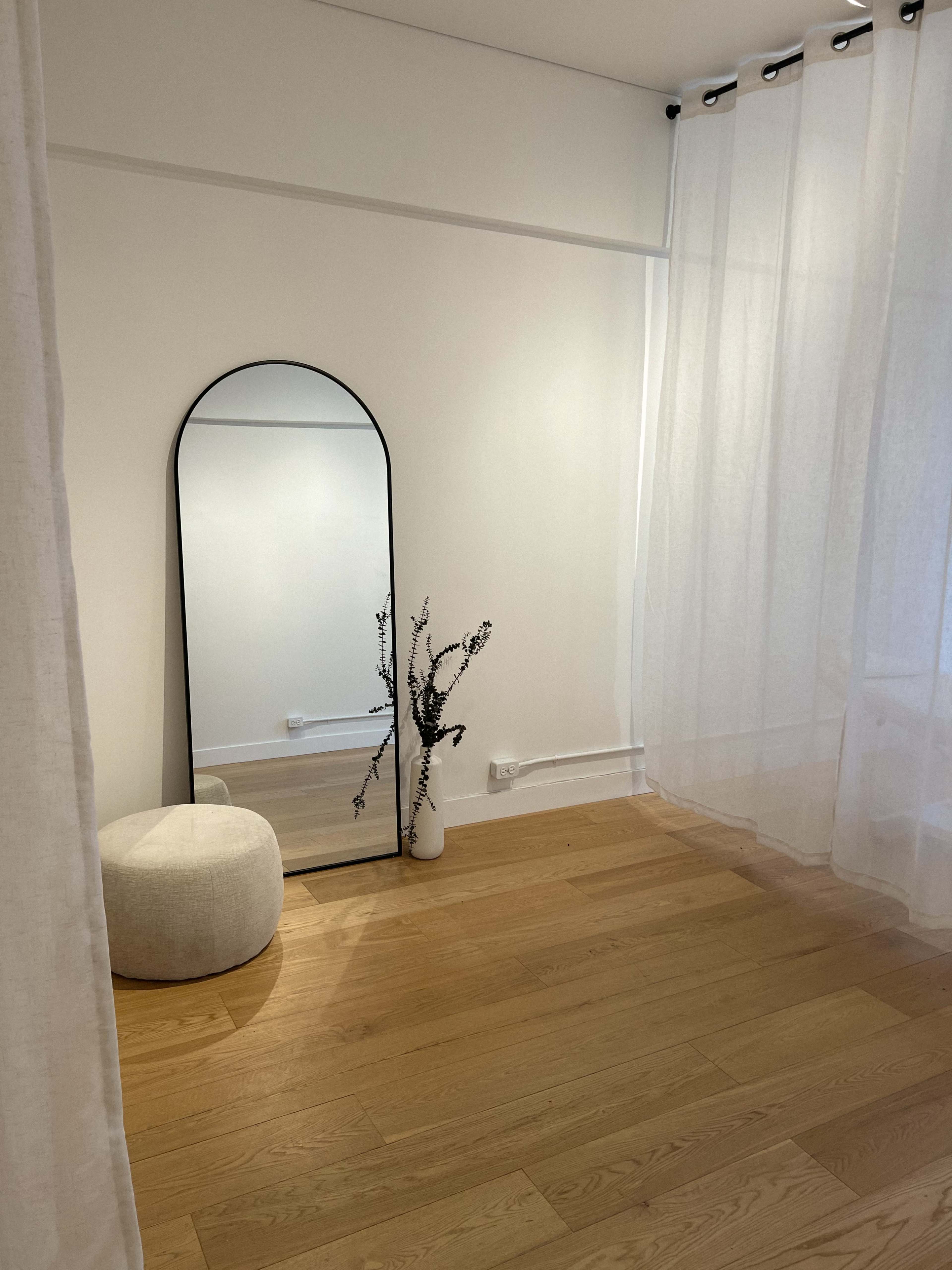 A minimalistic room features a large mirror, a white pouf, and a vase with branches beside sheer curtains and wooden flooring.