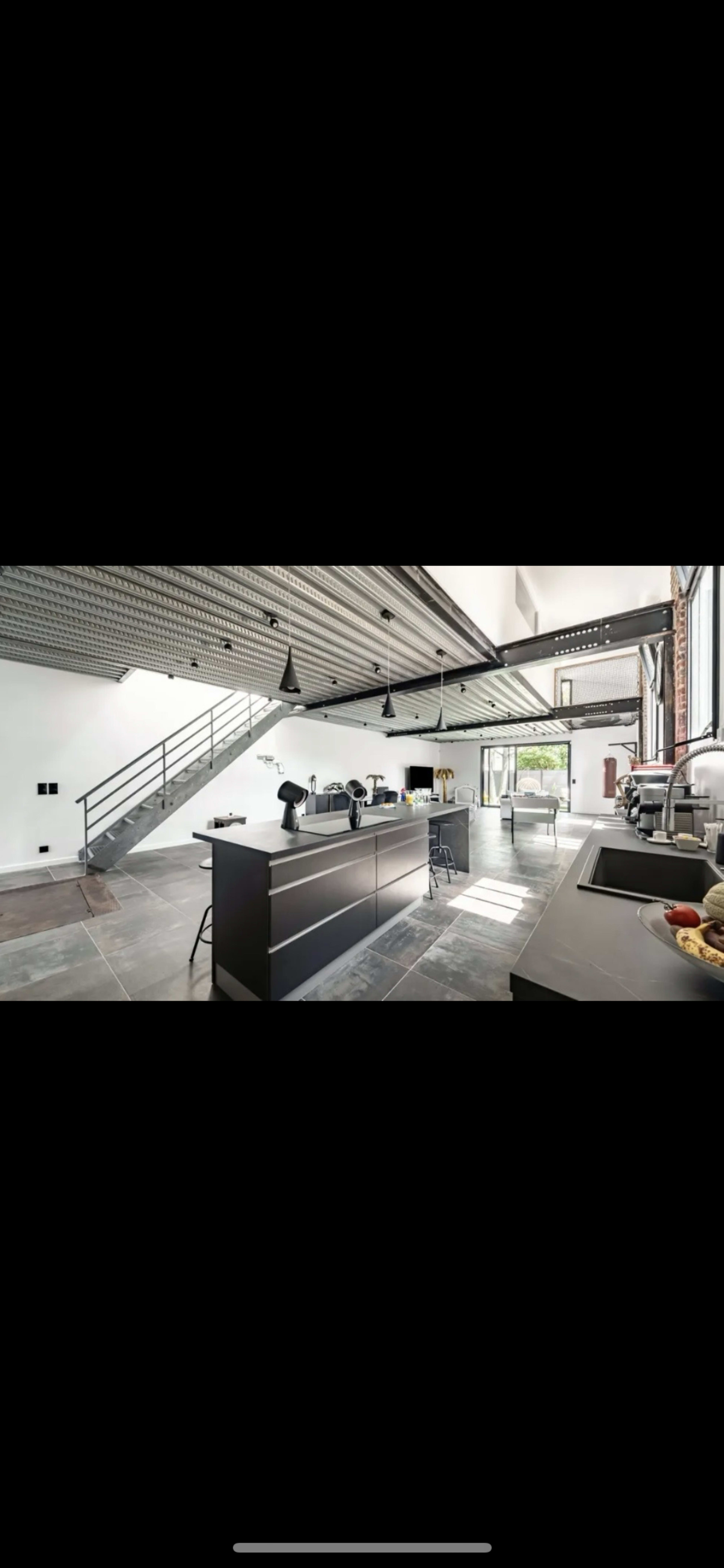 A modern, open-concept kitchen with gray cabinets and a large island, accompanied by a minimalist staircase and large windows allowing natural light to enter.