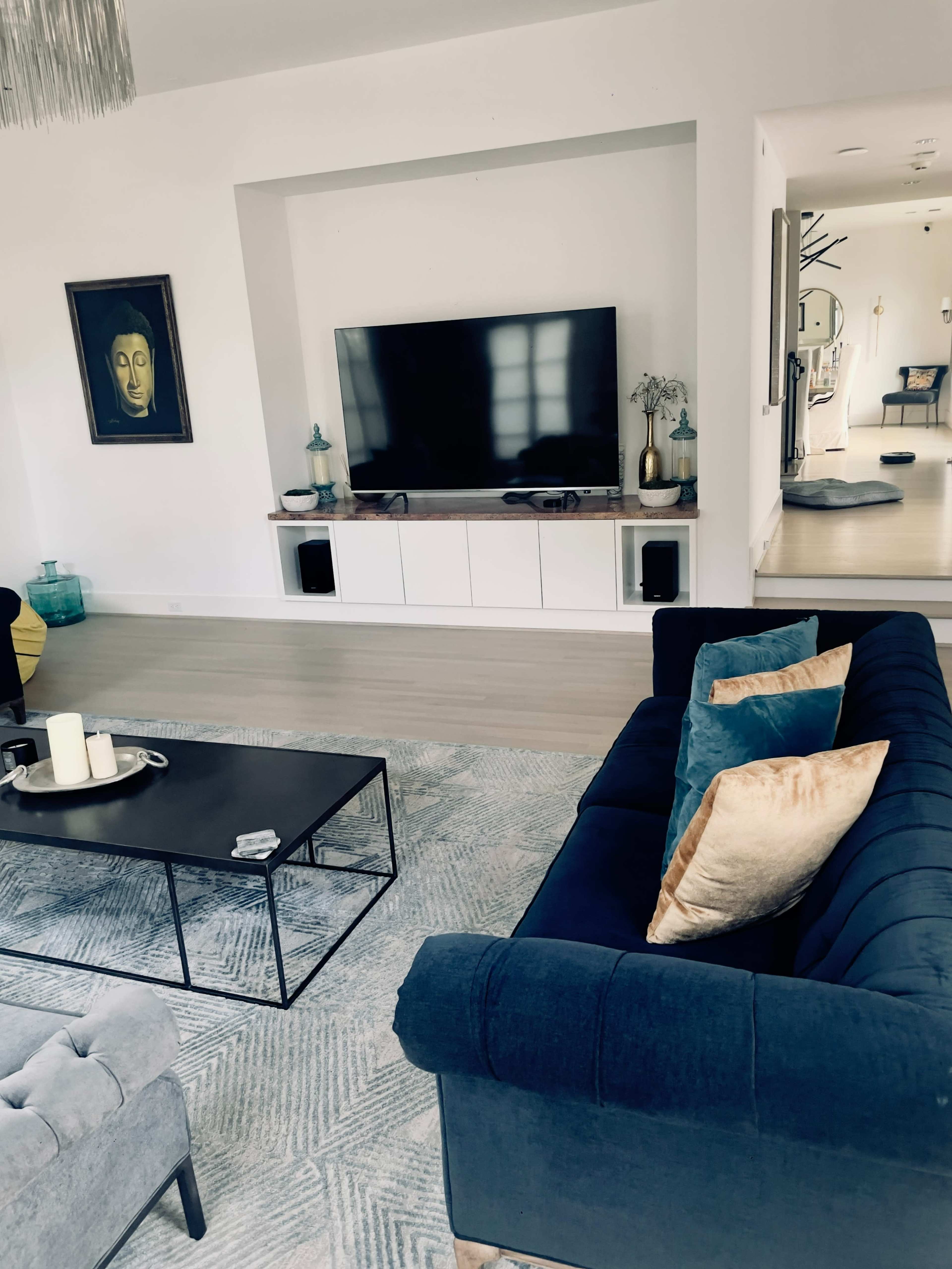 The image shows a modern living room with a large flat-screen TV mounted on a white wall, a black coffee table, and a blue couch with decorative cushions, all set against a light-colored floor.