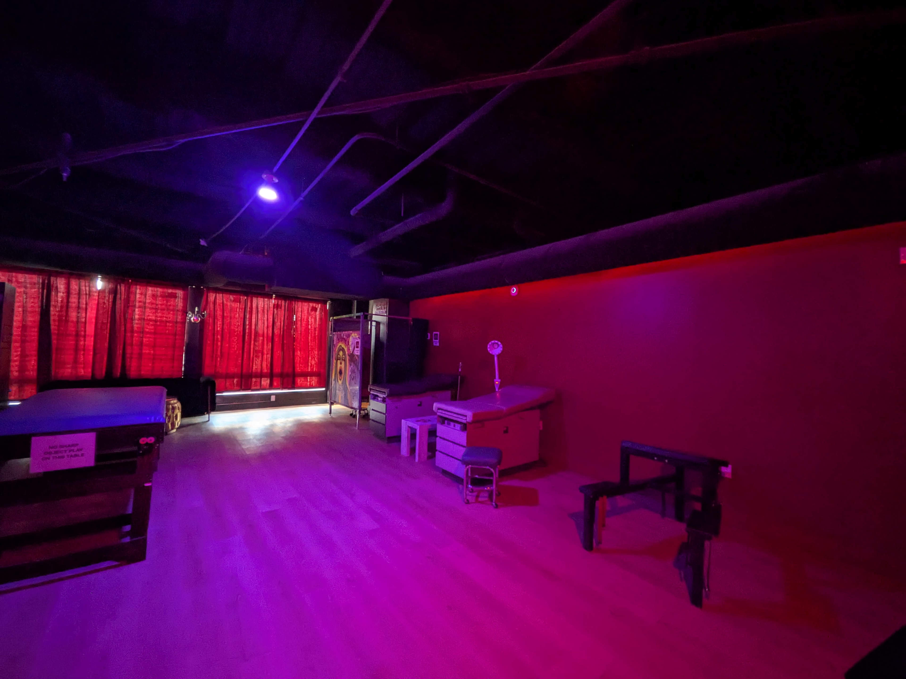 The room features a sparse layout with a dark ceiling, purple lighting, red curtains, and minimal furnishings including tables and a treatment bench.