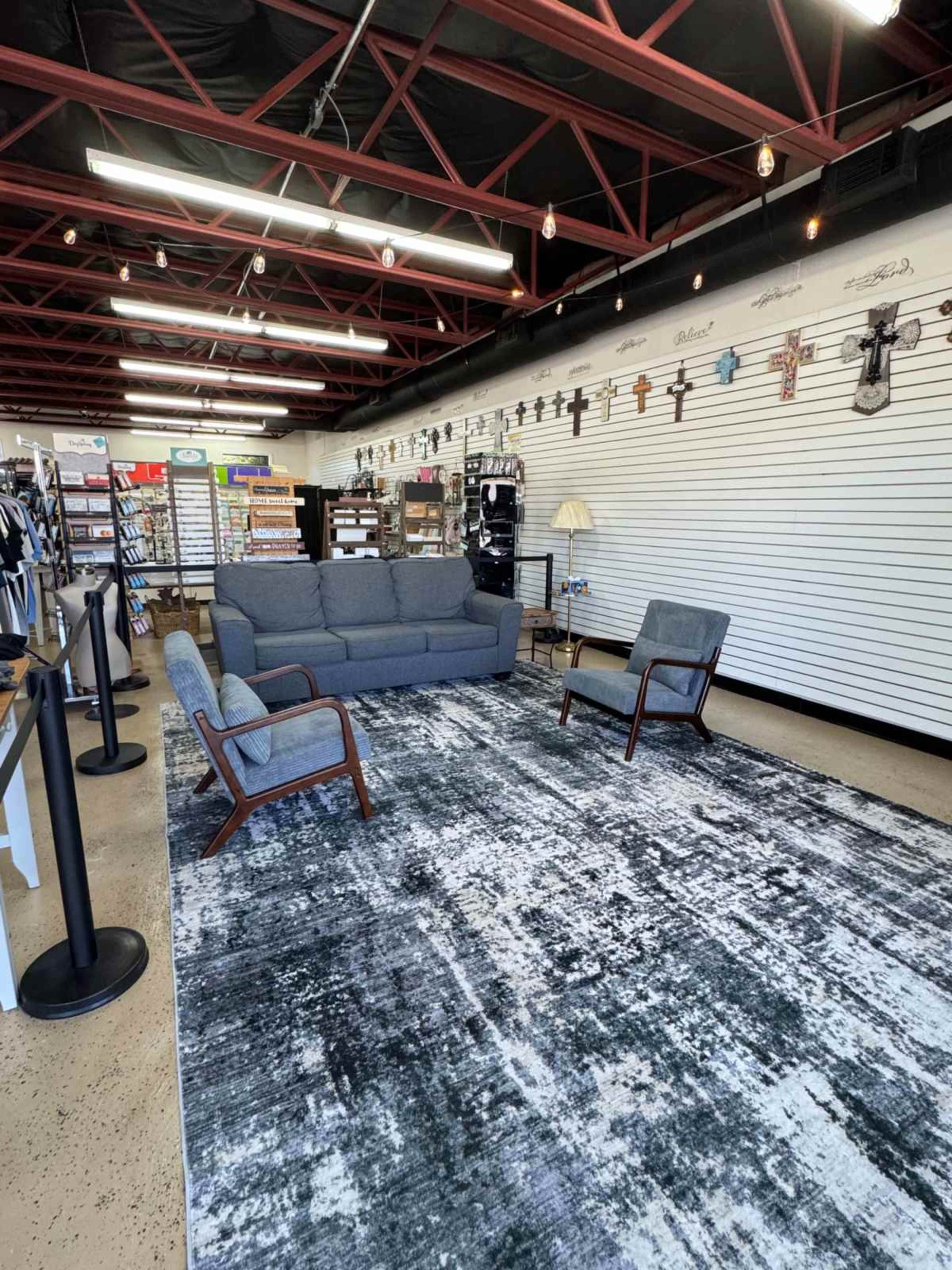 The image shows a seating area in a retail space featuring a gray sofa, two chairs, and a large area rug on a concrete floor, with merchandise displayed on slatwall behind.