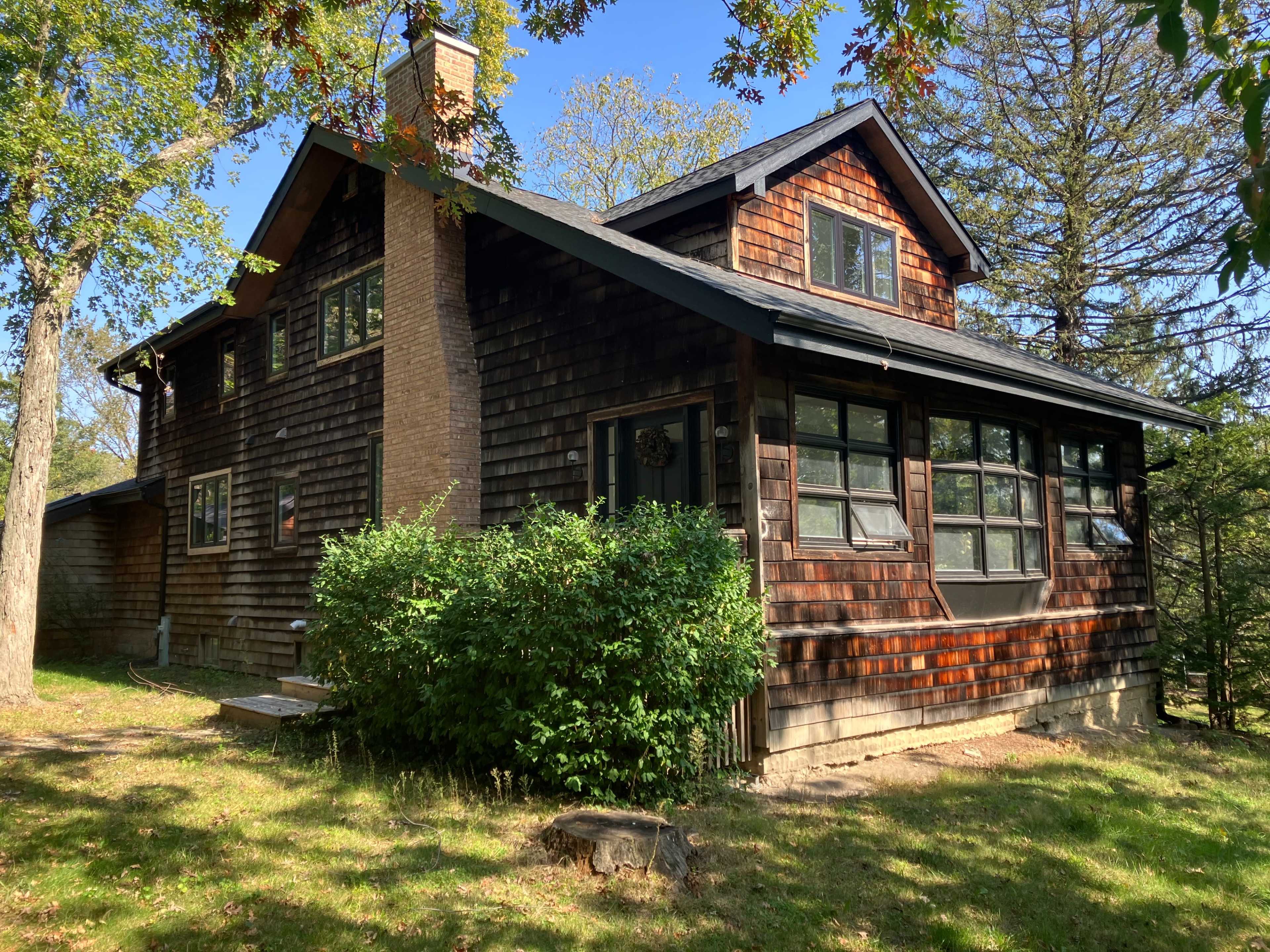The image shows a two-story wooden cabin with a stone chimney, surrounded by trees and grass.
