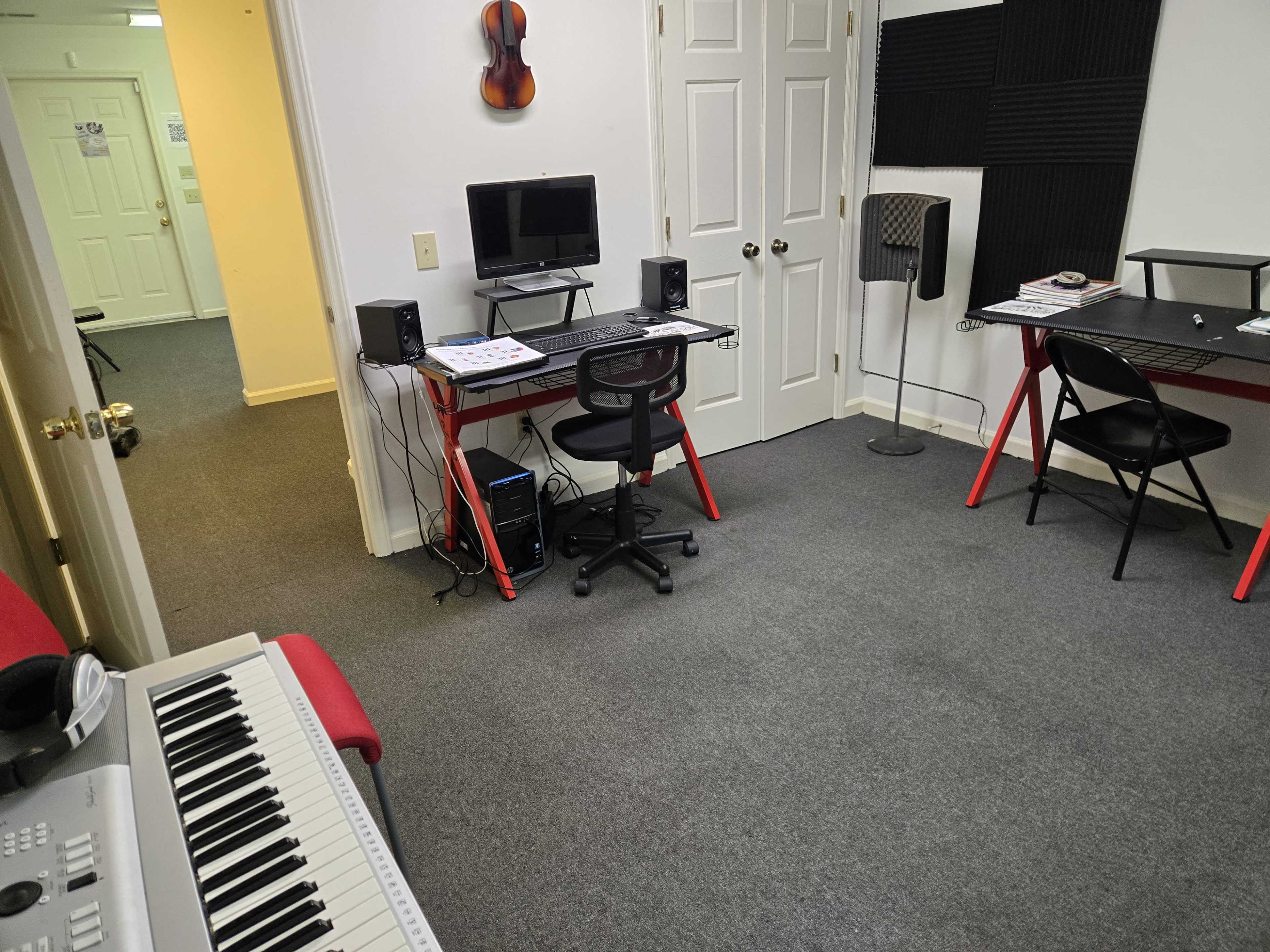 The image shows a small music studio with a computer desk, an electronic keyboard in the foreground, and a hallway leading to another room in the background.