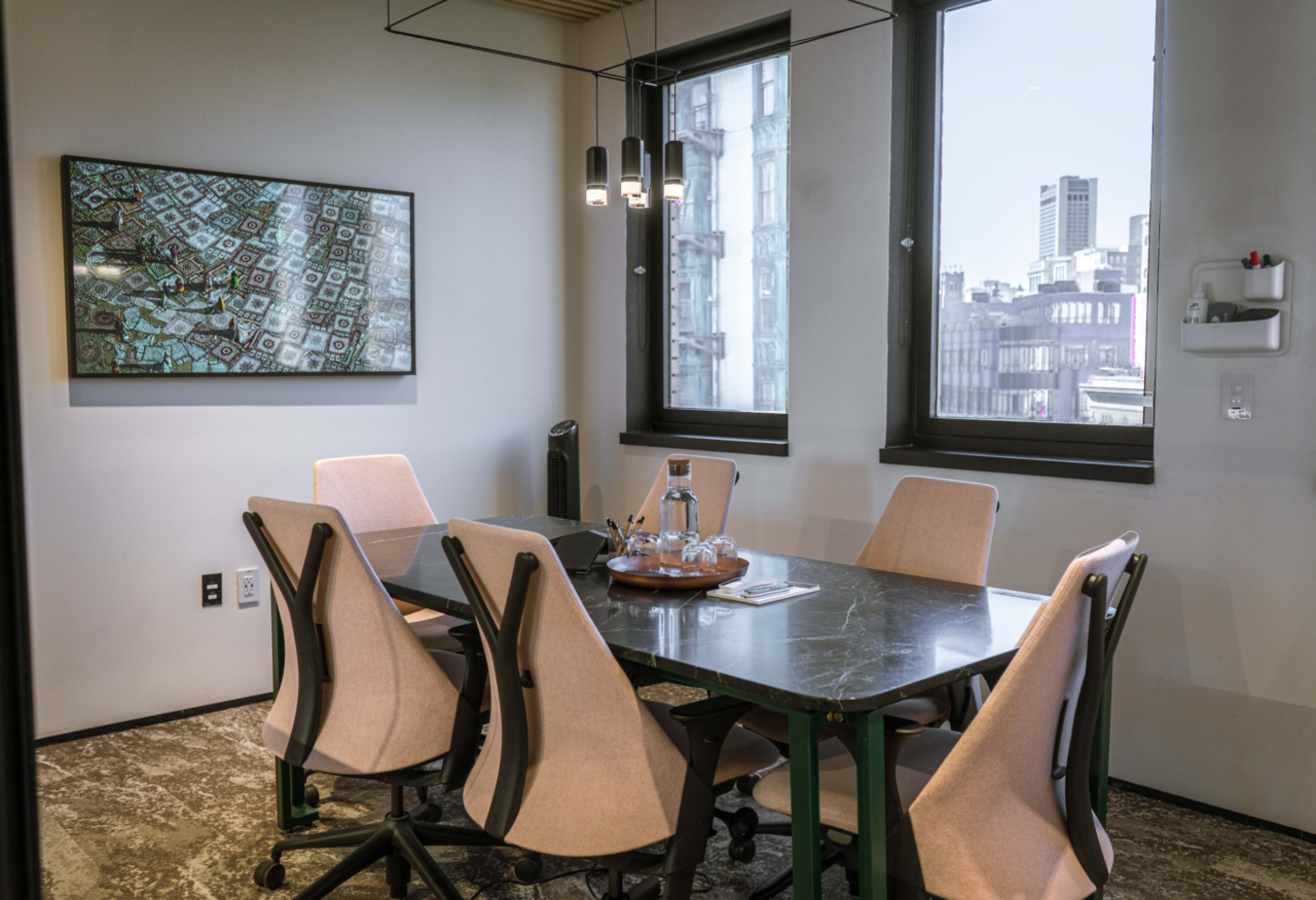 A modern conference room features a large table surrounded by six chairs, with a decorative painting on the wall and windows displaying a cityscape.