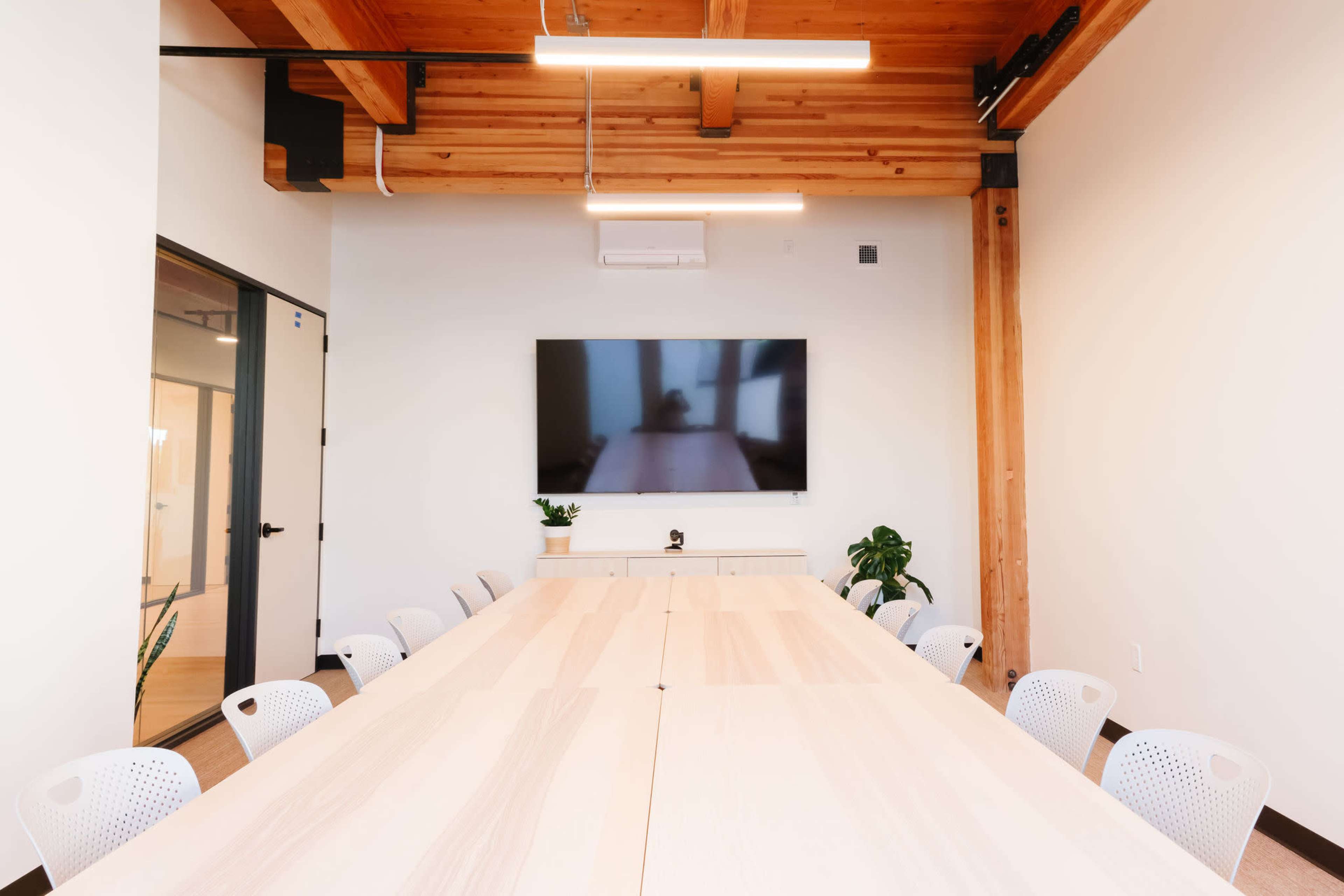 A conference room features a long wooden table surrounded by chairs, with a large wall-mounted screen and plants in the corners.