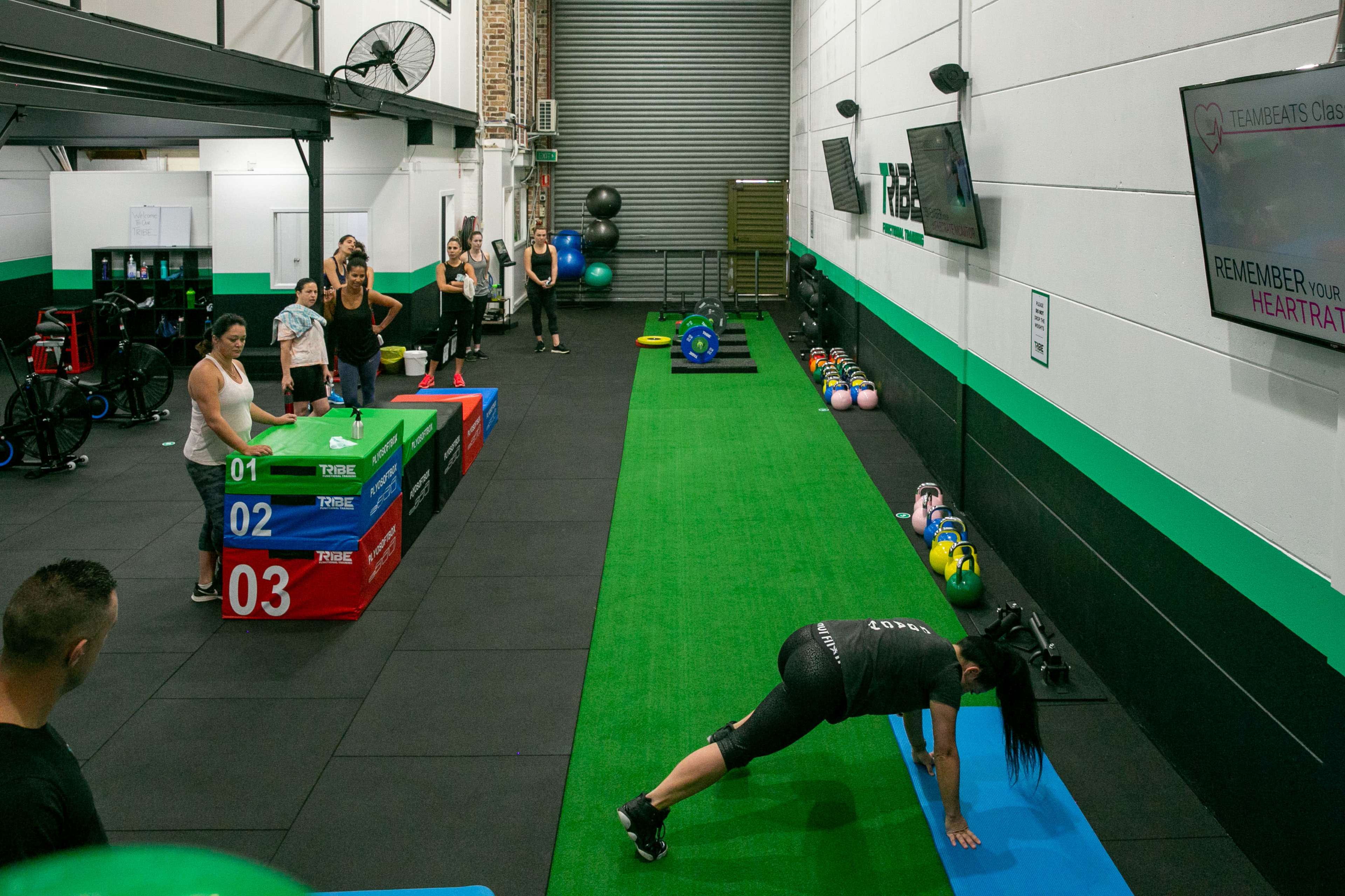 A fitness class is taking place in a gym, with participants exercising on colorful platforms and equipment while an instructor guides them.