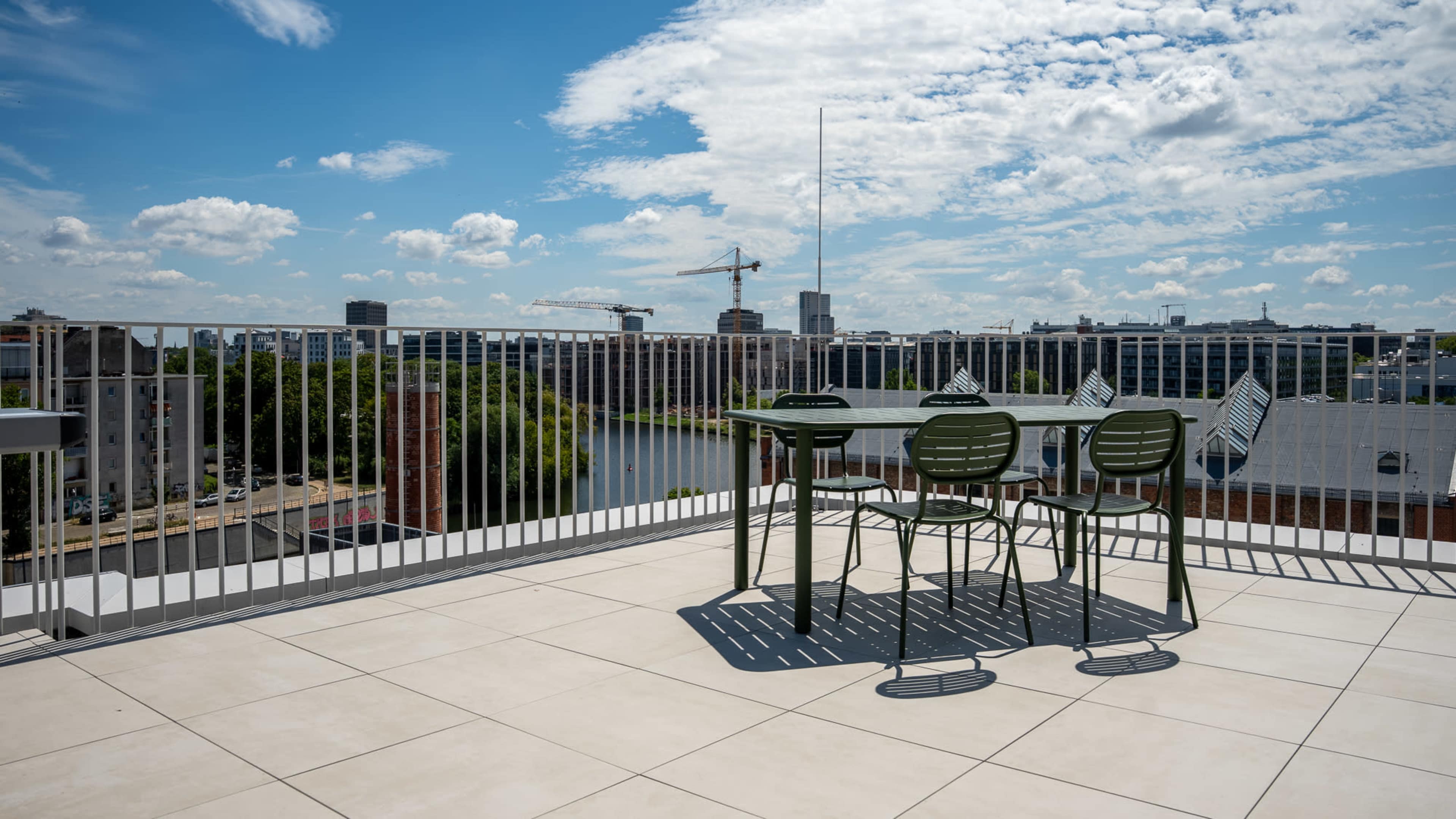 A rooftop terrace features a table and chairs with a view of a city skyline and a river under a partly cloudy sky.