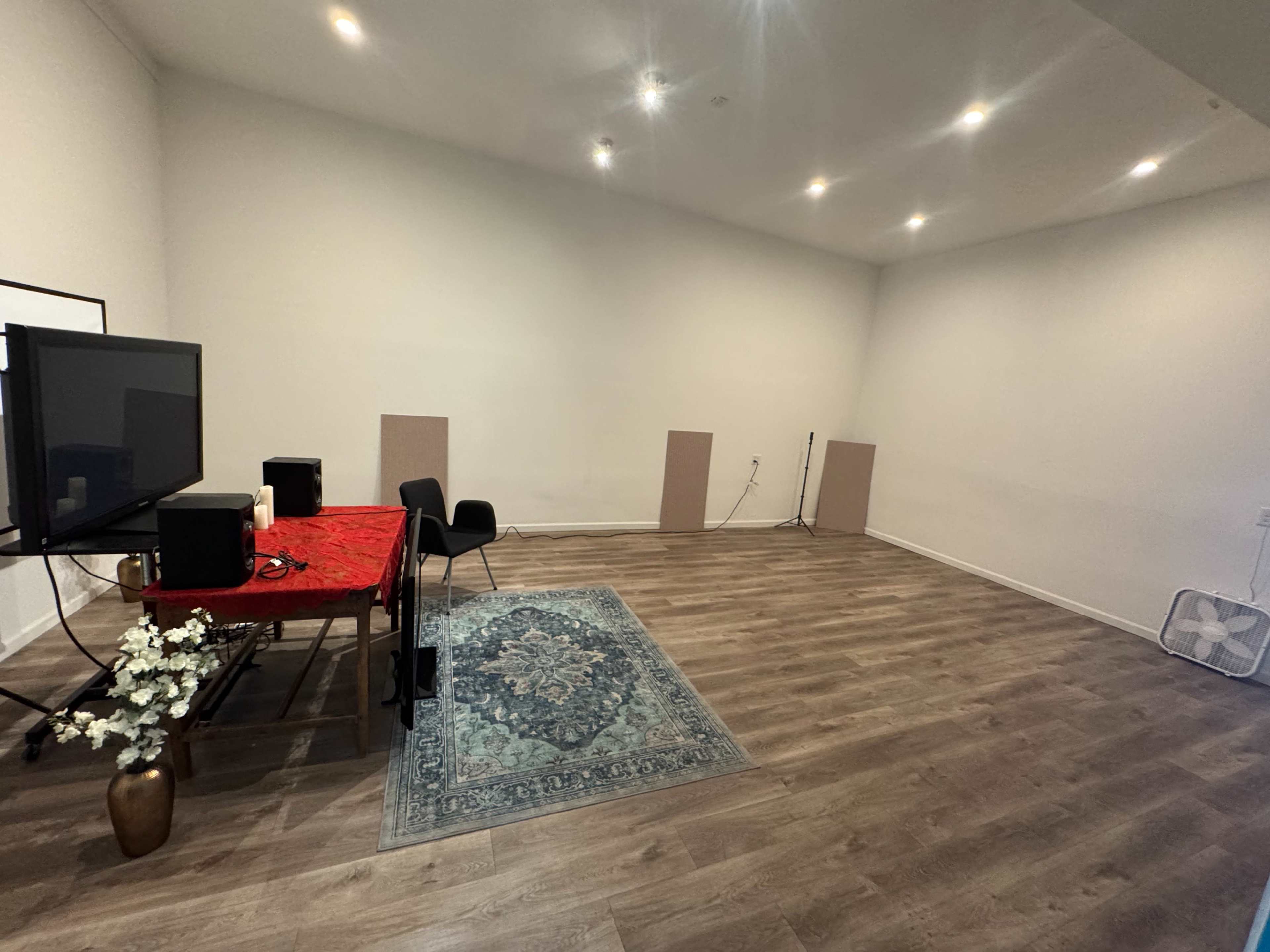 The image shows a spacious room with wooden flooring, a decorative rug, a table with a red tablecloth, a television, speakers, and a fan.