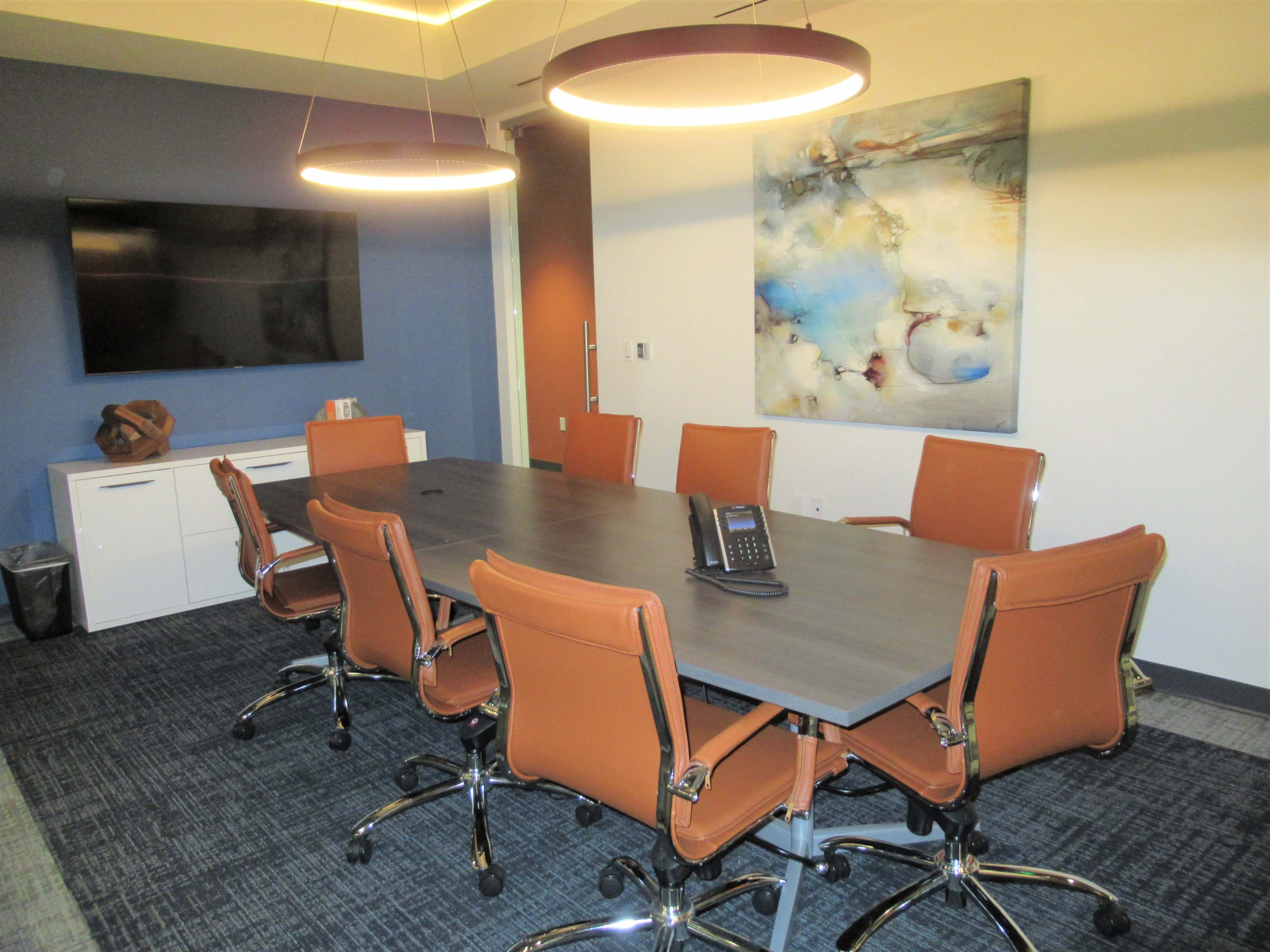 A modern conference room with a large rectangular table surrounded by eight orange office chairs, a phone located on the table, and a television mounted on the wall.