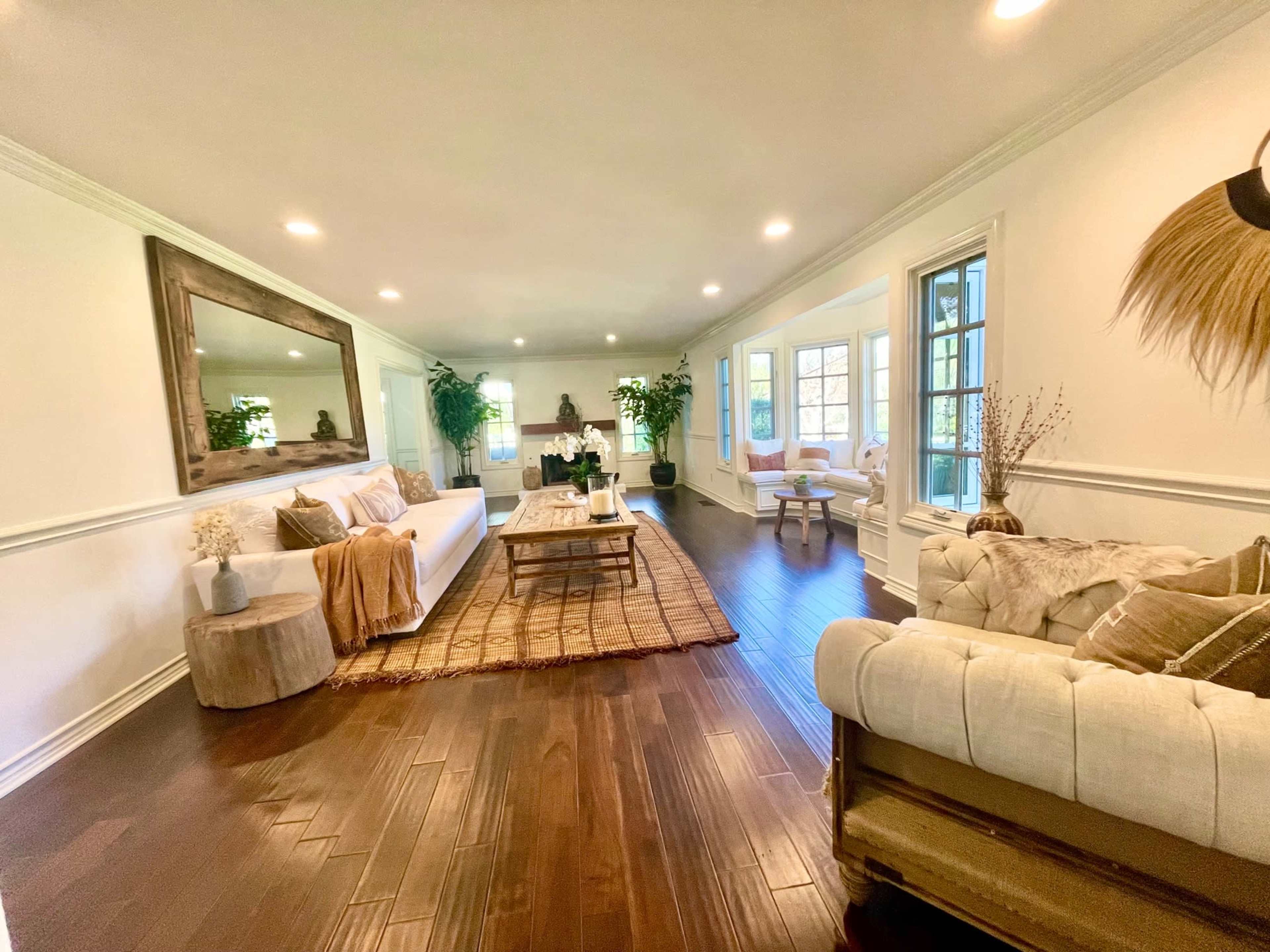 The spacious living room features a white sofa, wooden coffee table, and a mix of plants, with large windows allowing natural light to enter.