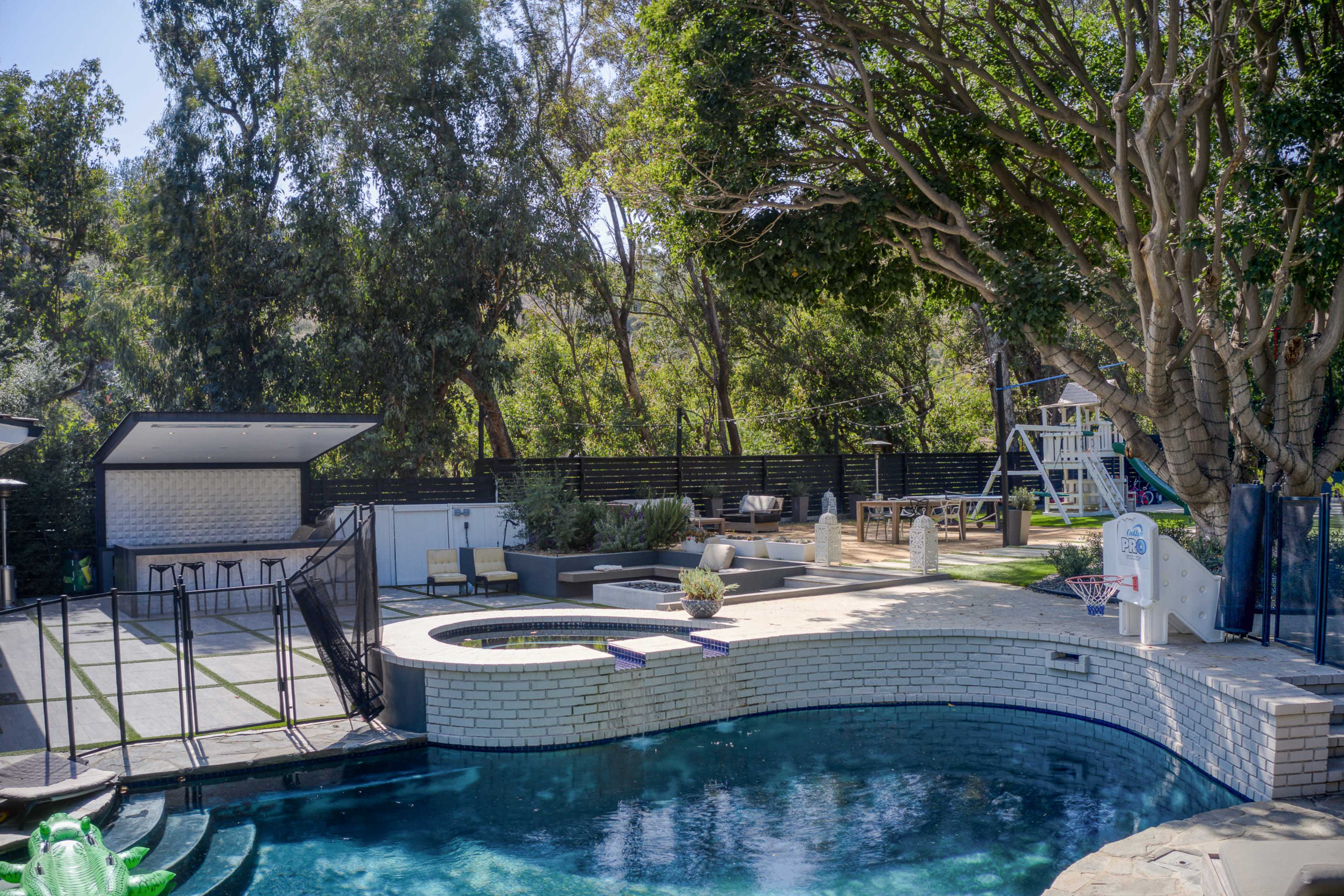 The image depicts a backyard with a swimming pool, surrounded by trees, lounge chairs, and a small outdoor dining area.