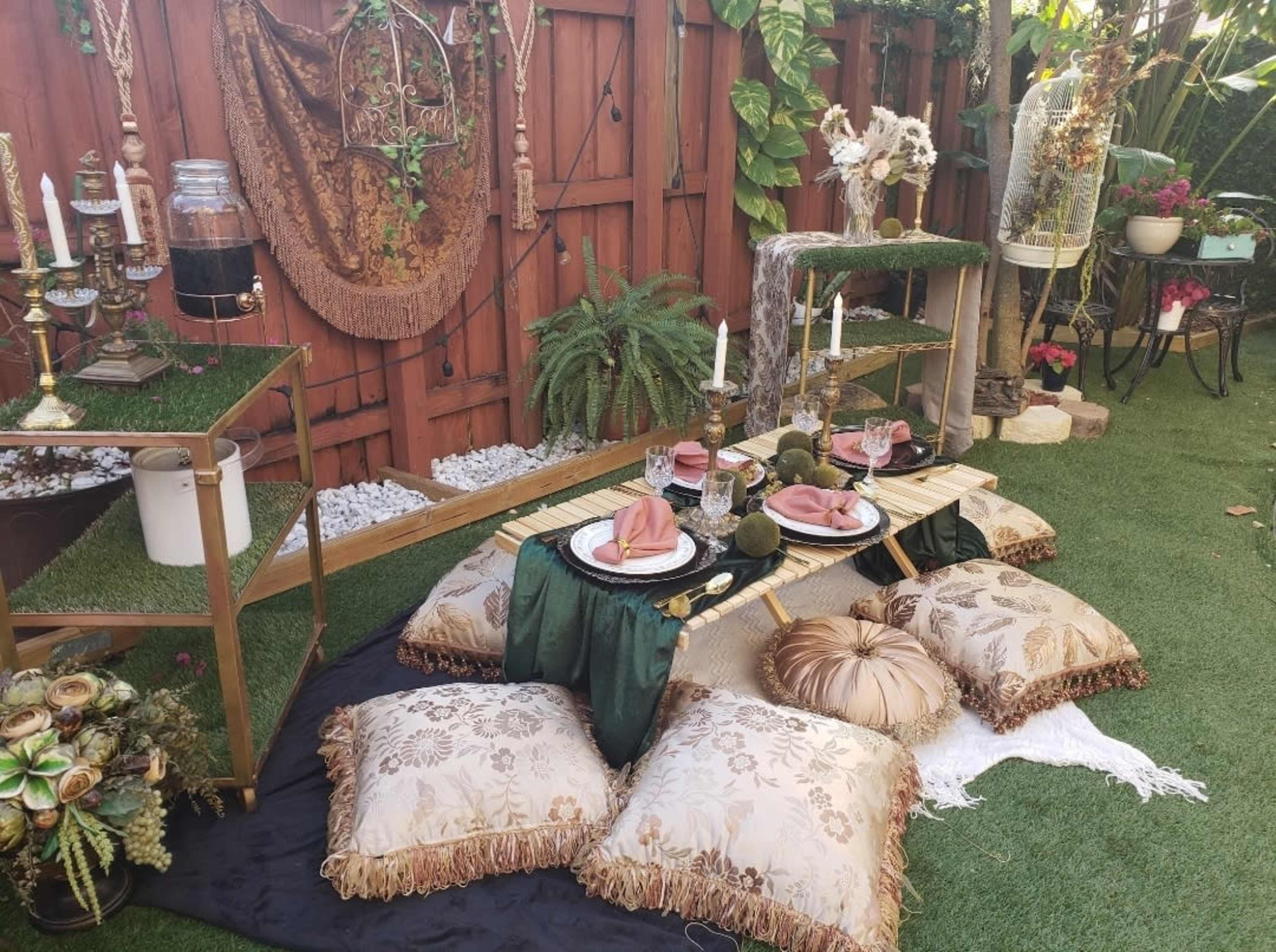 A low dining setup features a bamboo table adorned with dishes and cutlery, surrounded by decorative pillows on a grassy area against a wooden fence.