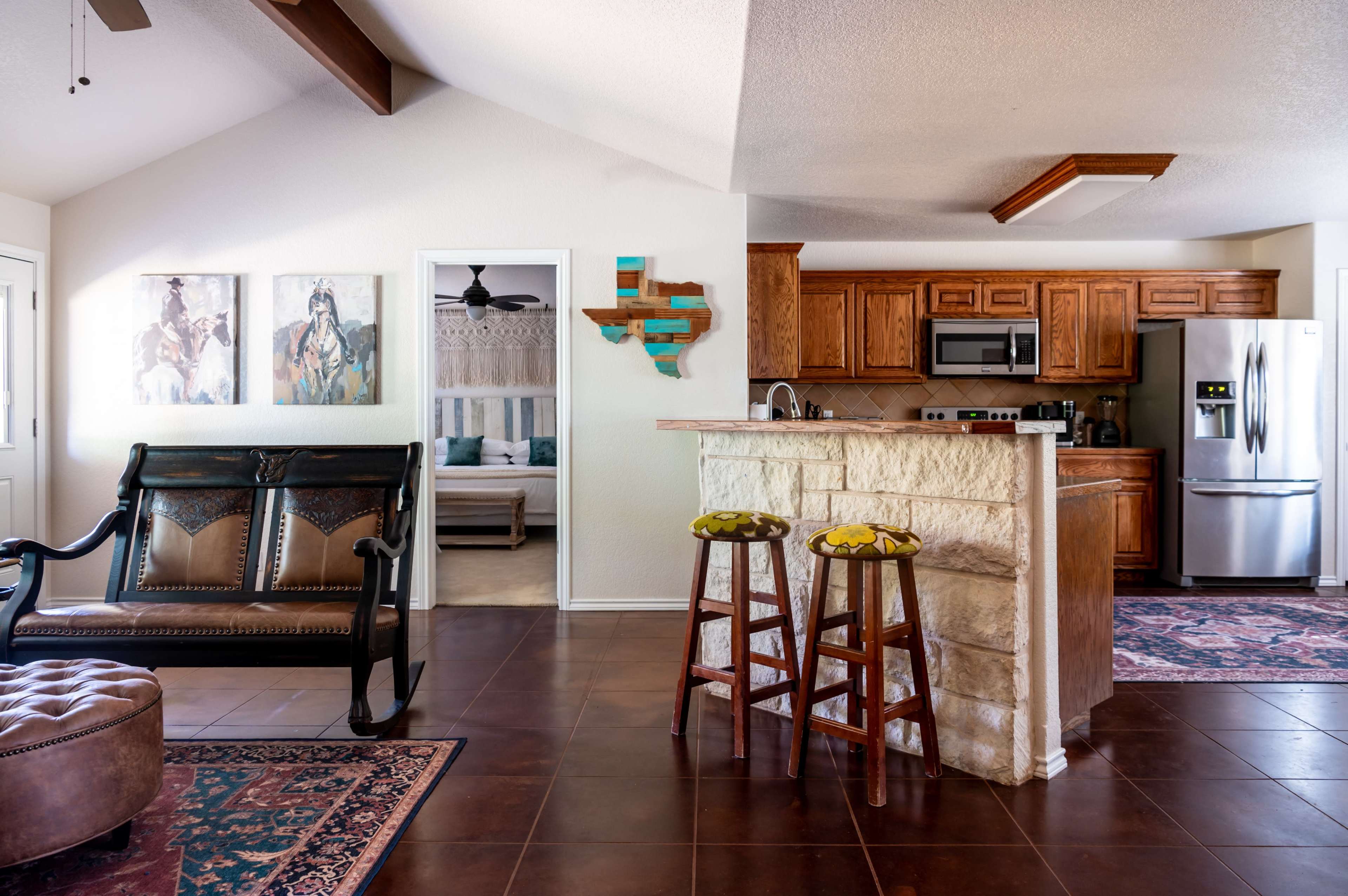 The image shows a spacious open-concept living area with a kitchen, featuring wooden cabinetry, a stone bar, and seating areas along with decorative wall art.