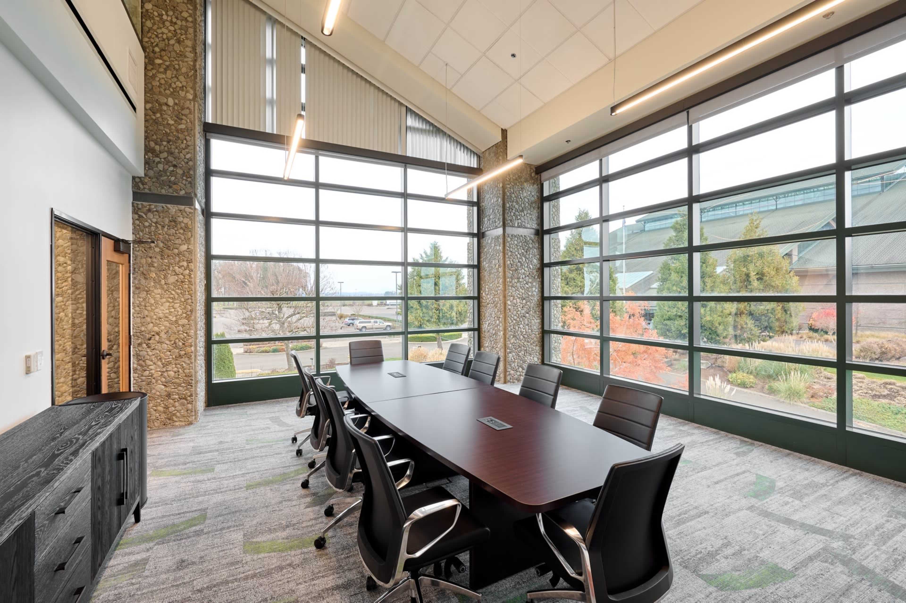The image shows a modern conference room with a large rectangular table surrounded by black chairs and large windows offering a view of greenery outside.