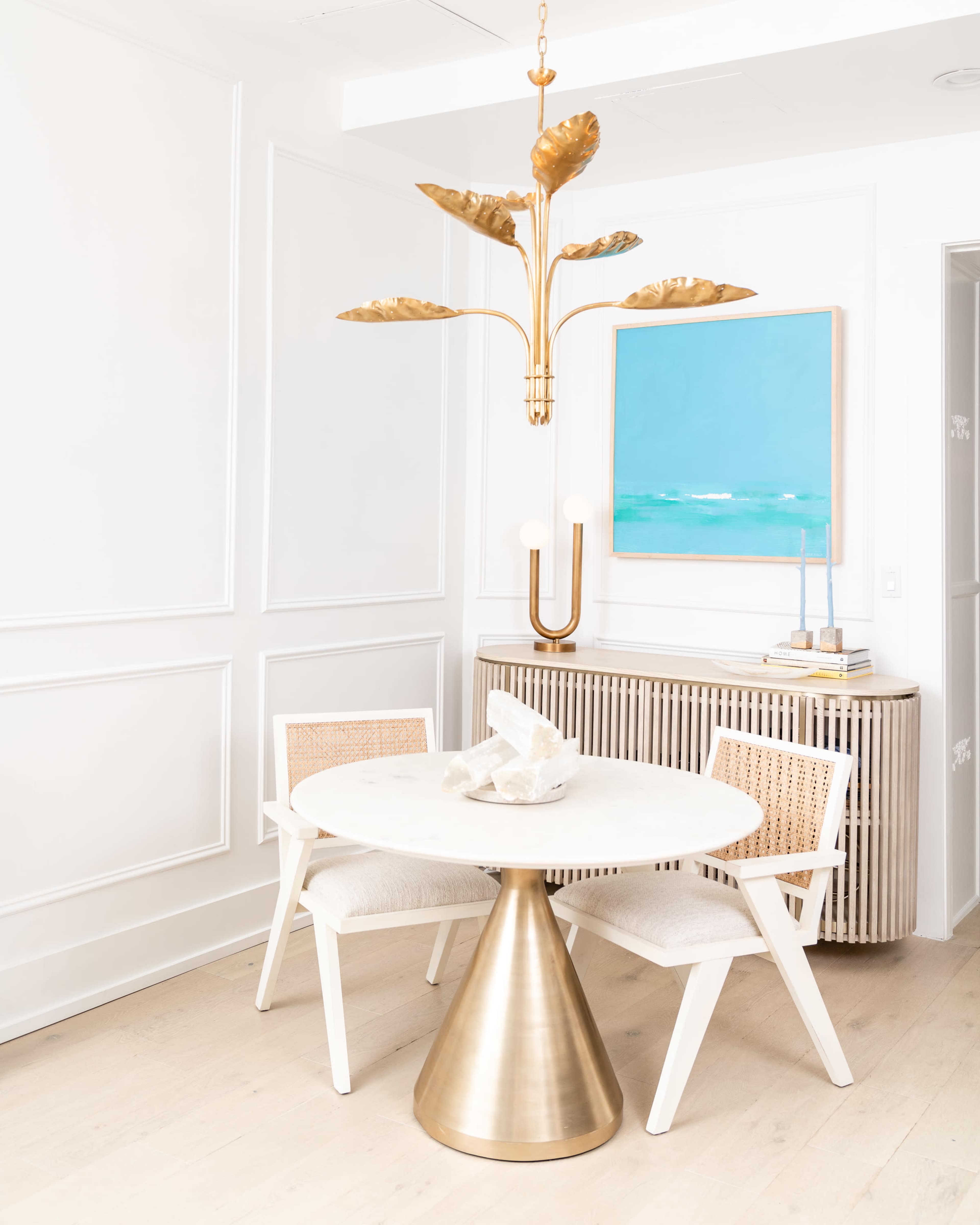 A round marble dining table with a brass base is placed between two white chairs, beneath a gold leaf chandelier, in a room featuring a light blue wall art and a striped sideboard.