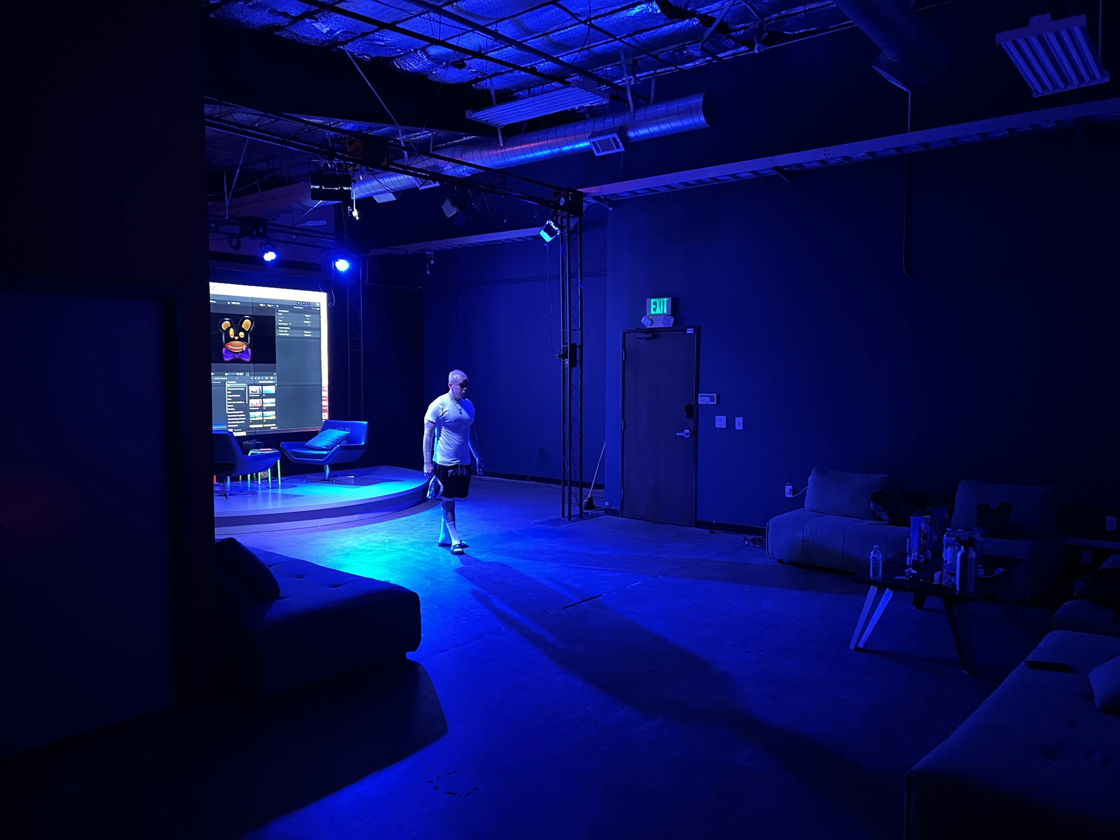 A person walks through a dark room illuminated by blue lights, with seating arrangements and a large screen displaying information in the background.