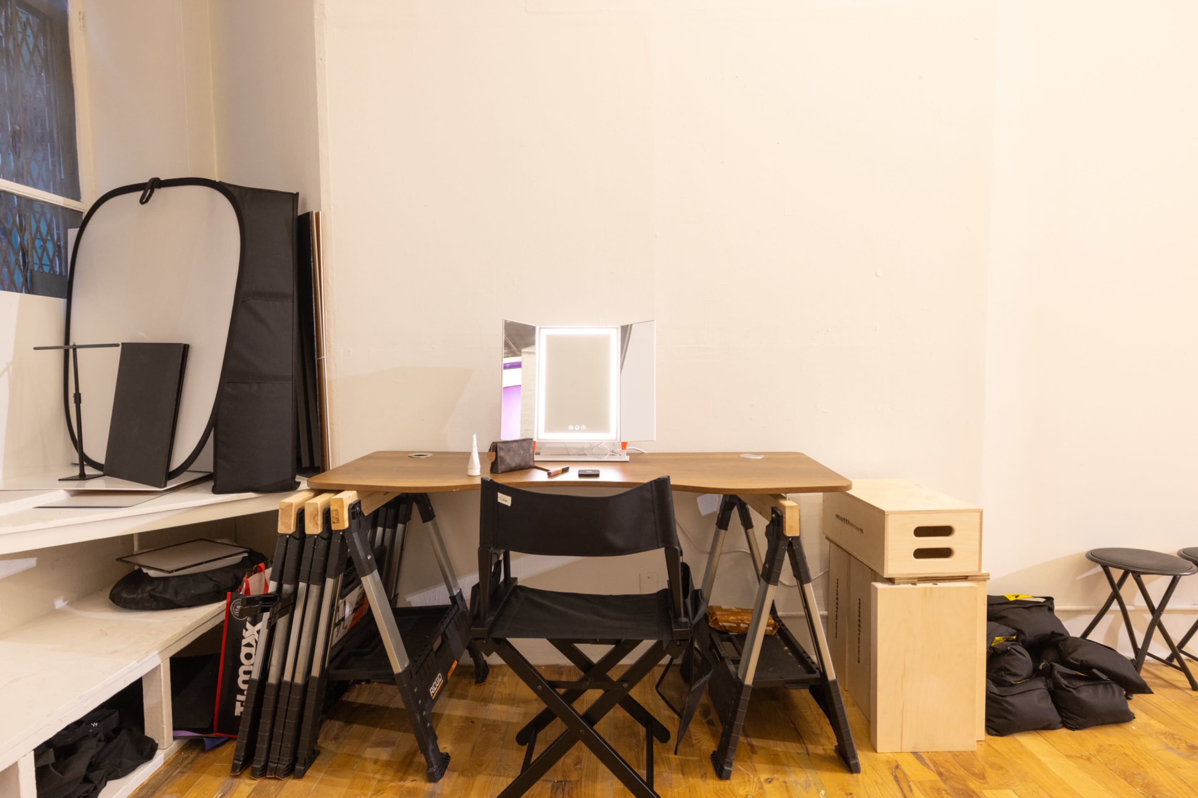 The image shows a minimalist workspace with a wooden desk, a mirror, and various storage boxes, surrounded by folding chairs and photography equipment.