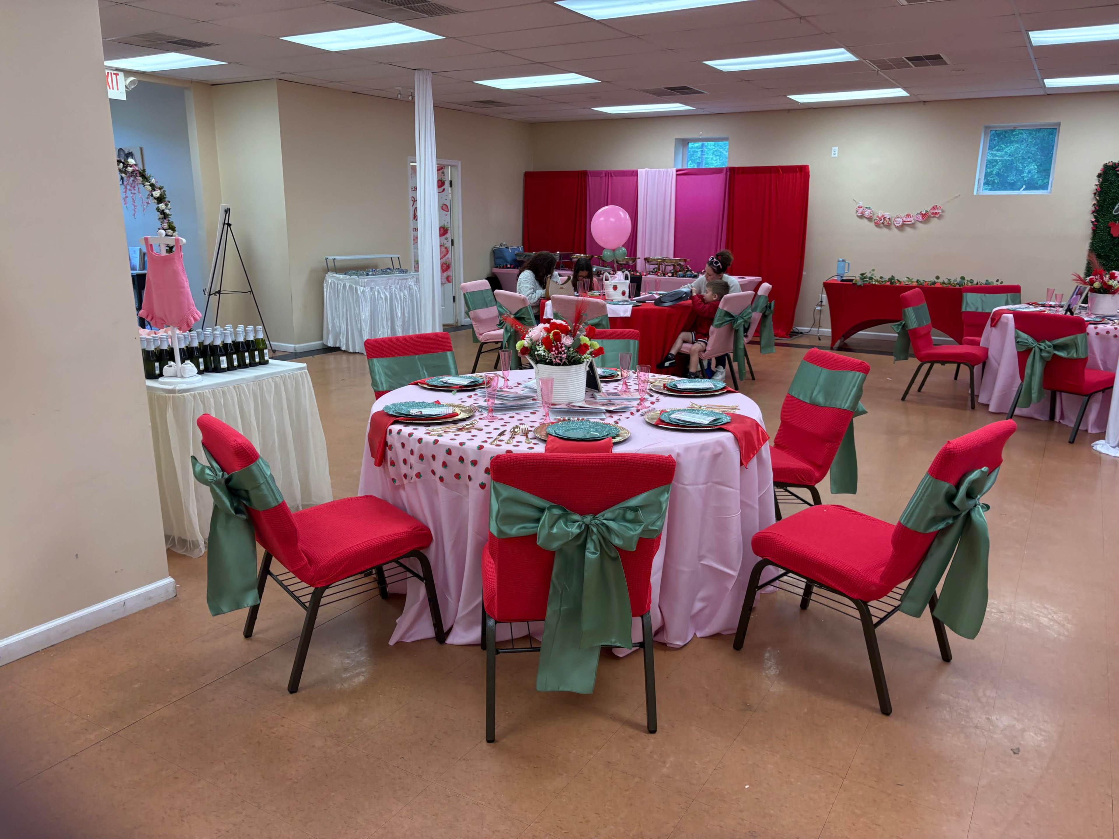 A decorated event space features red and white tables with floral centerpieces, pink balloons, and guests seated at tables.