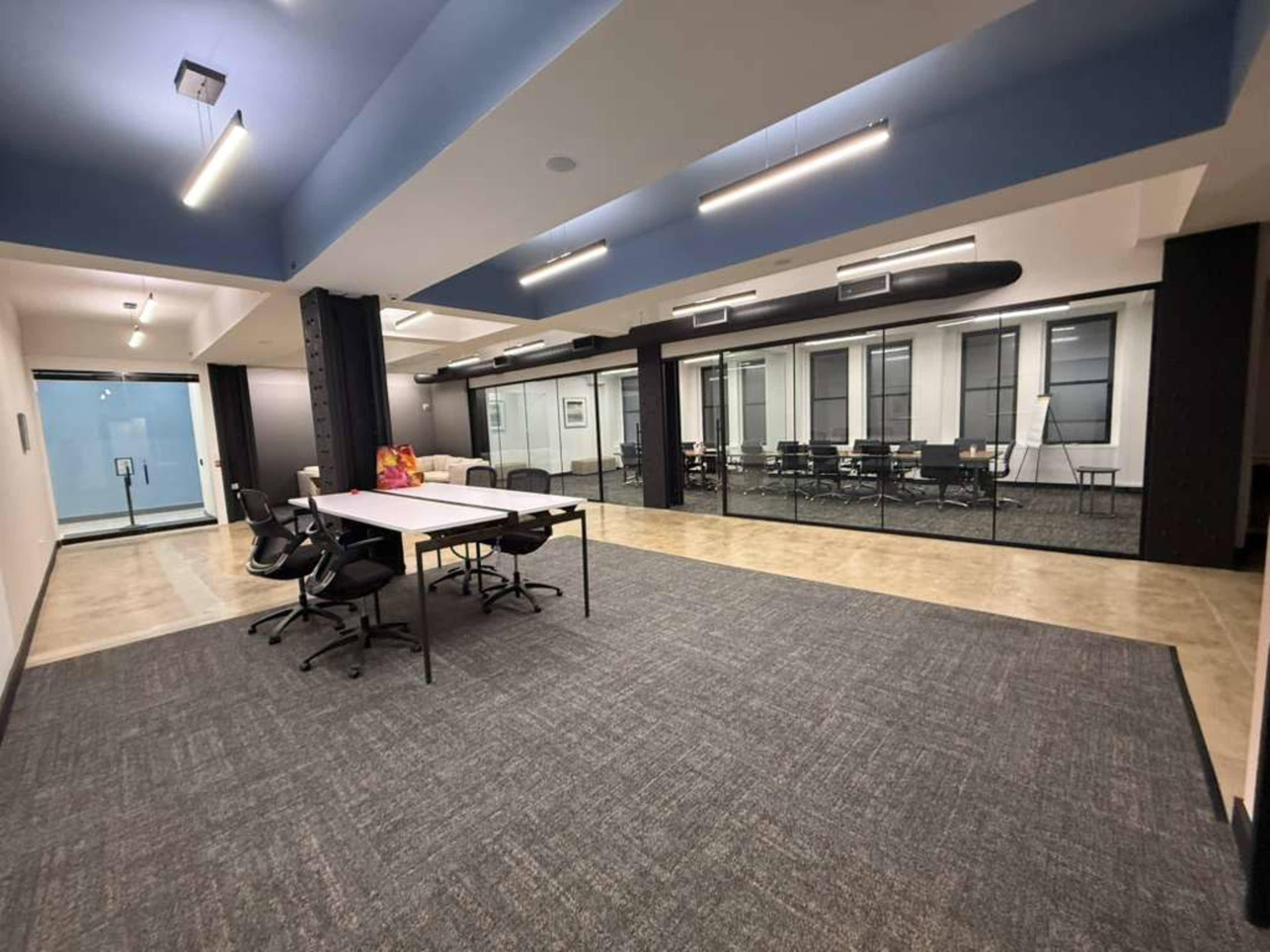 The image shows a modern office space featuring a central table with chairs, surrounded by glass-enclosed meeting rooms.