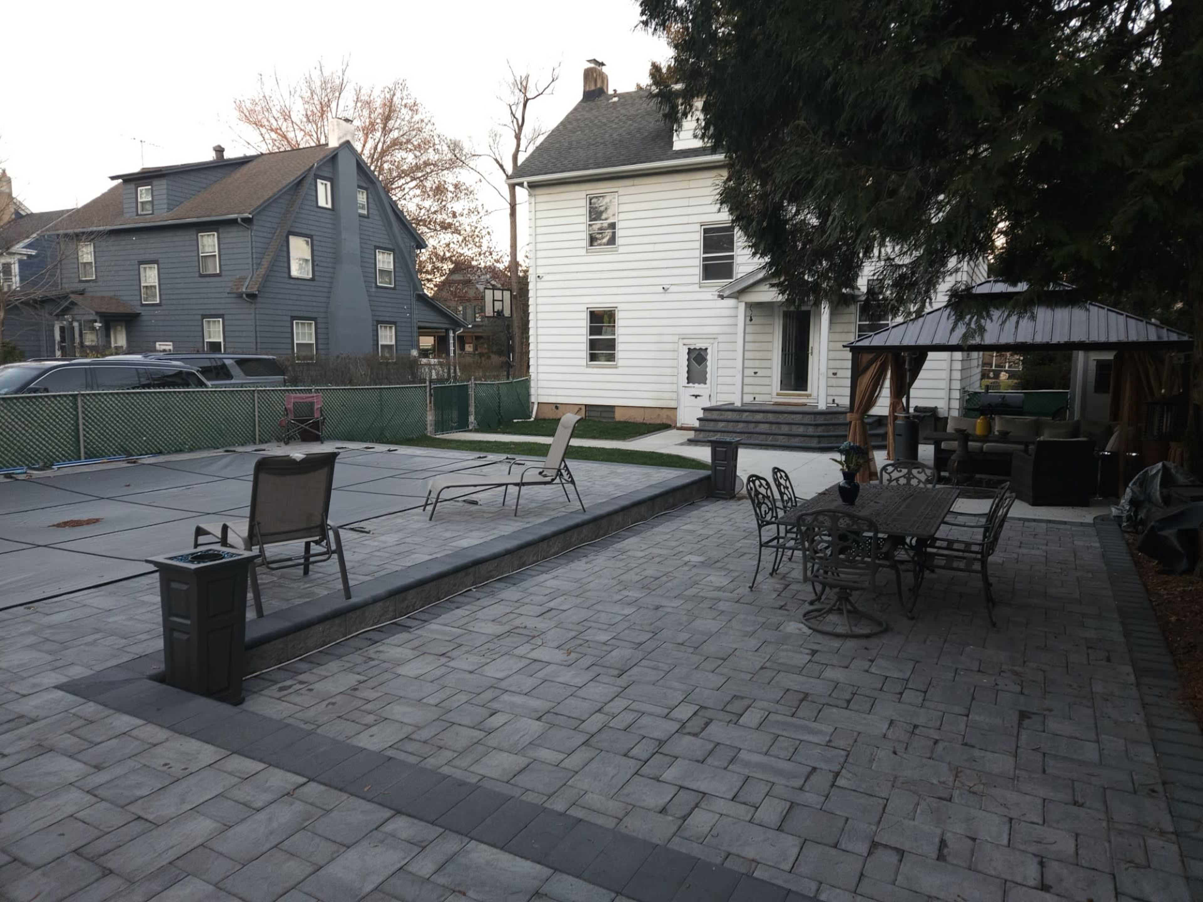 A backyard patio area with a pool covered and surrounded by paver stones, featuring lounge chairs, a dining set, and a gazebo.