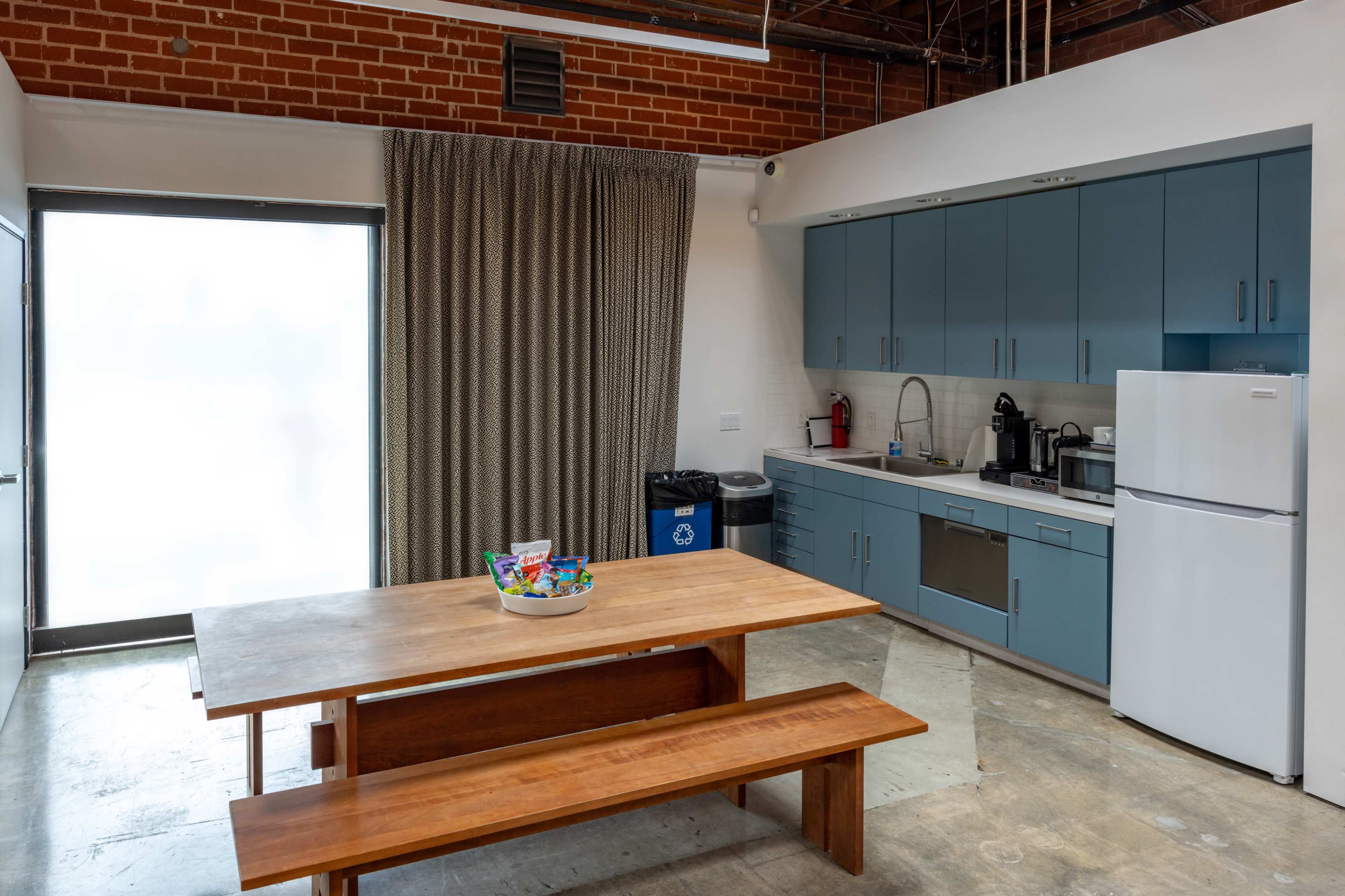 The image shows a modern kitchen with blue cabinetry, a wooden dining table, and a window covered by a curtain.