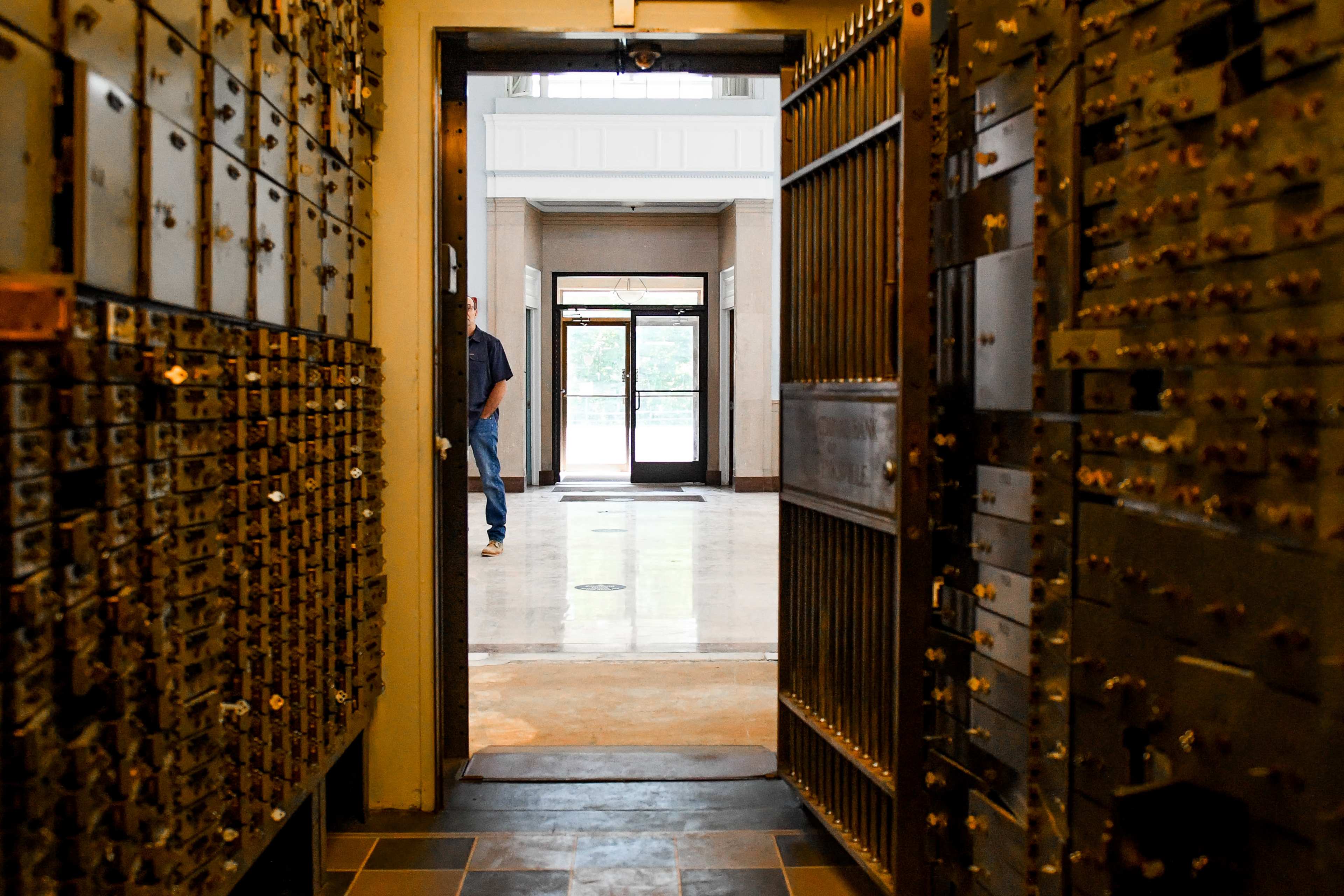 The image shows the interior of a bank vault with numerous safety deposit boxes on the left, leading to a doorway that opens into a brightly lit hallway.