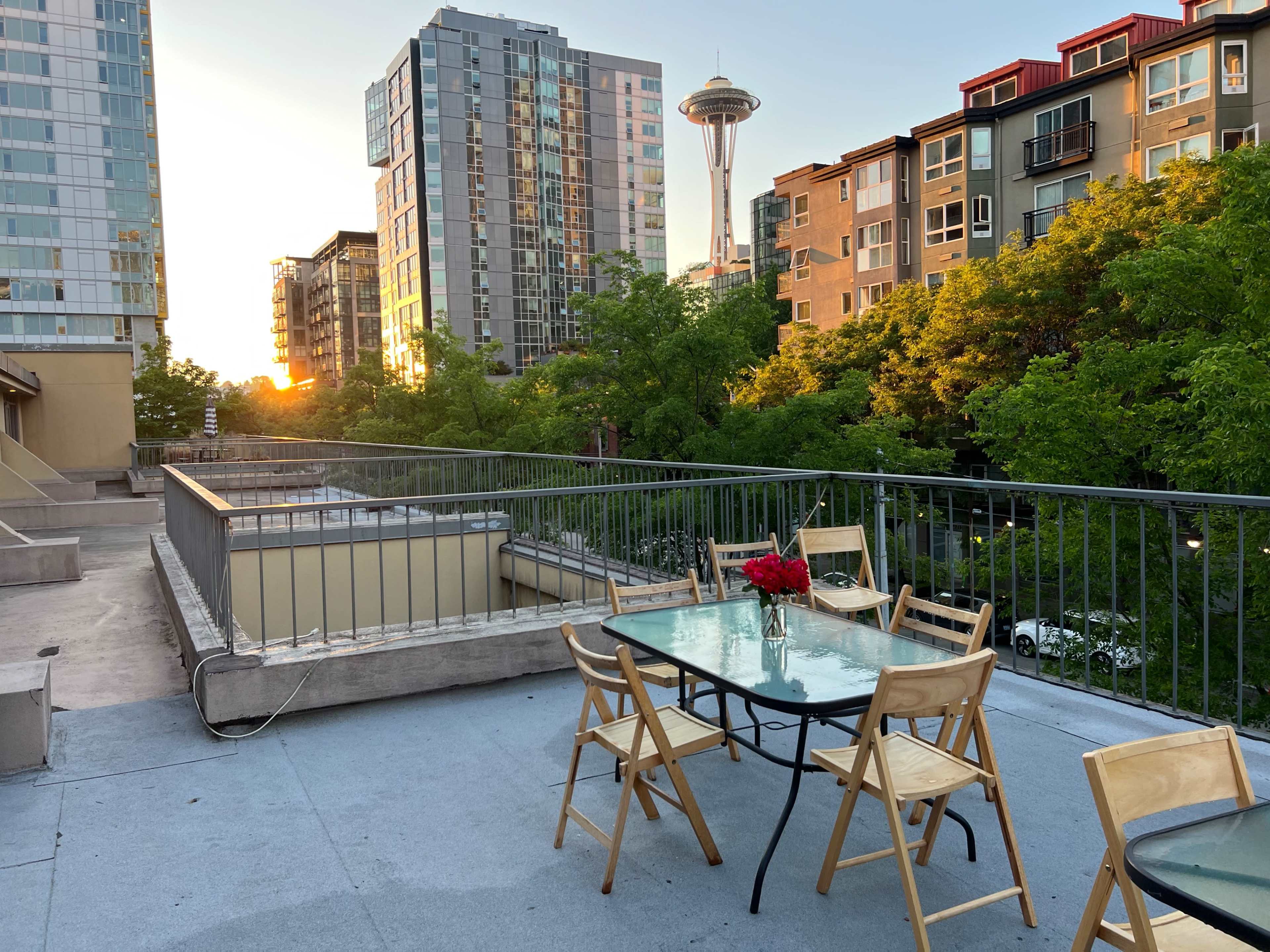 A rooftop patio with a glass table and wooden chairs overlooks a cityscape at sunset, featuring the Space Needle in the background.