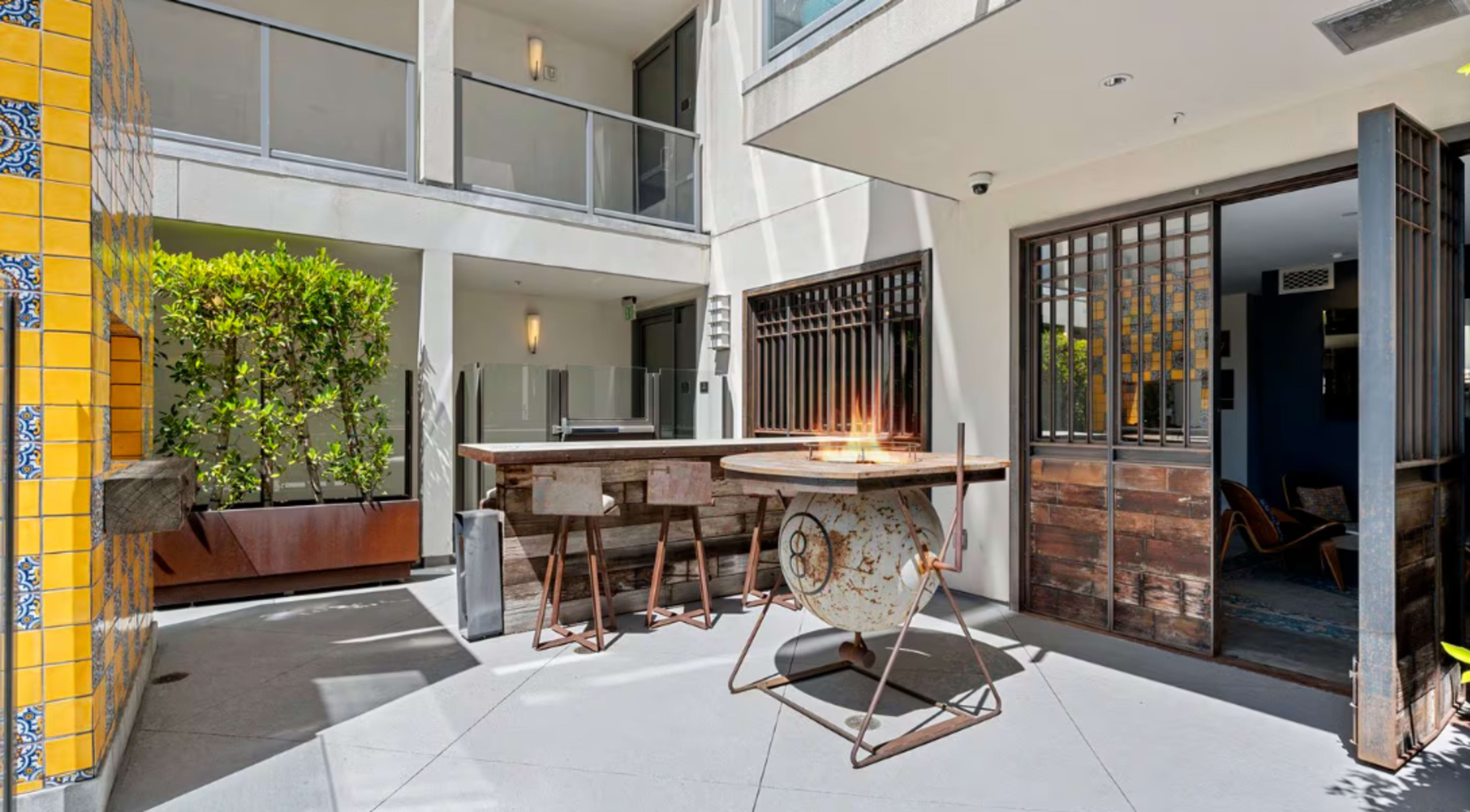 The image shows a modern courtyard with a bar area featuring a round table and stools, surrounded by greenery and architectural details.